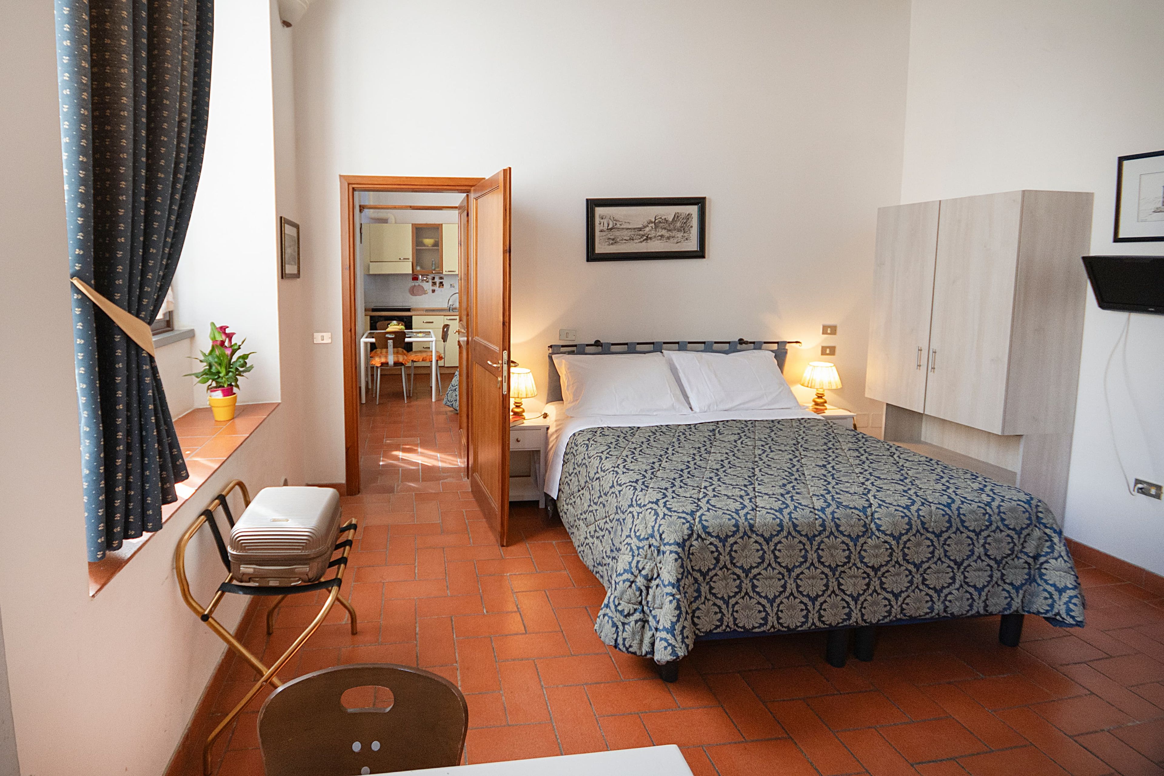 A bedroom with a bed, bedside table, lamp, and a view of a kitchen and bathroom through an open door.