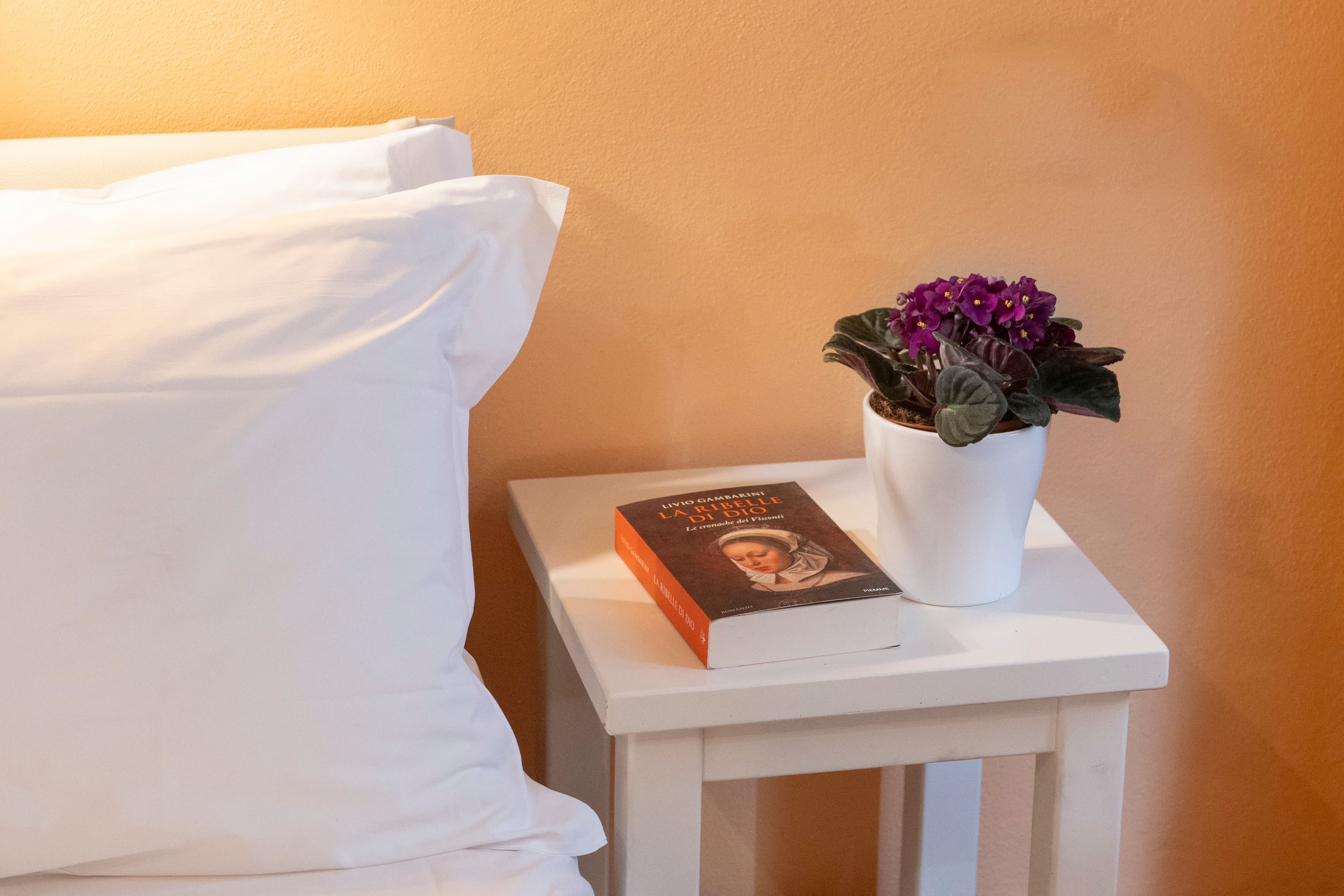 A white bedside table with a book titled 'La Rivibella di Dio' and a potted plant with purple flowers.