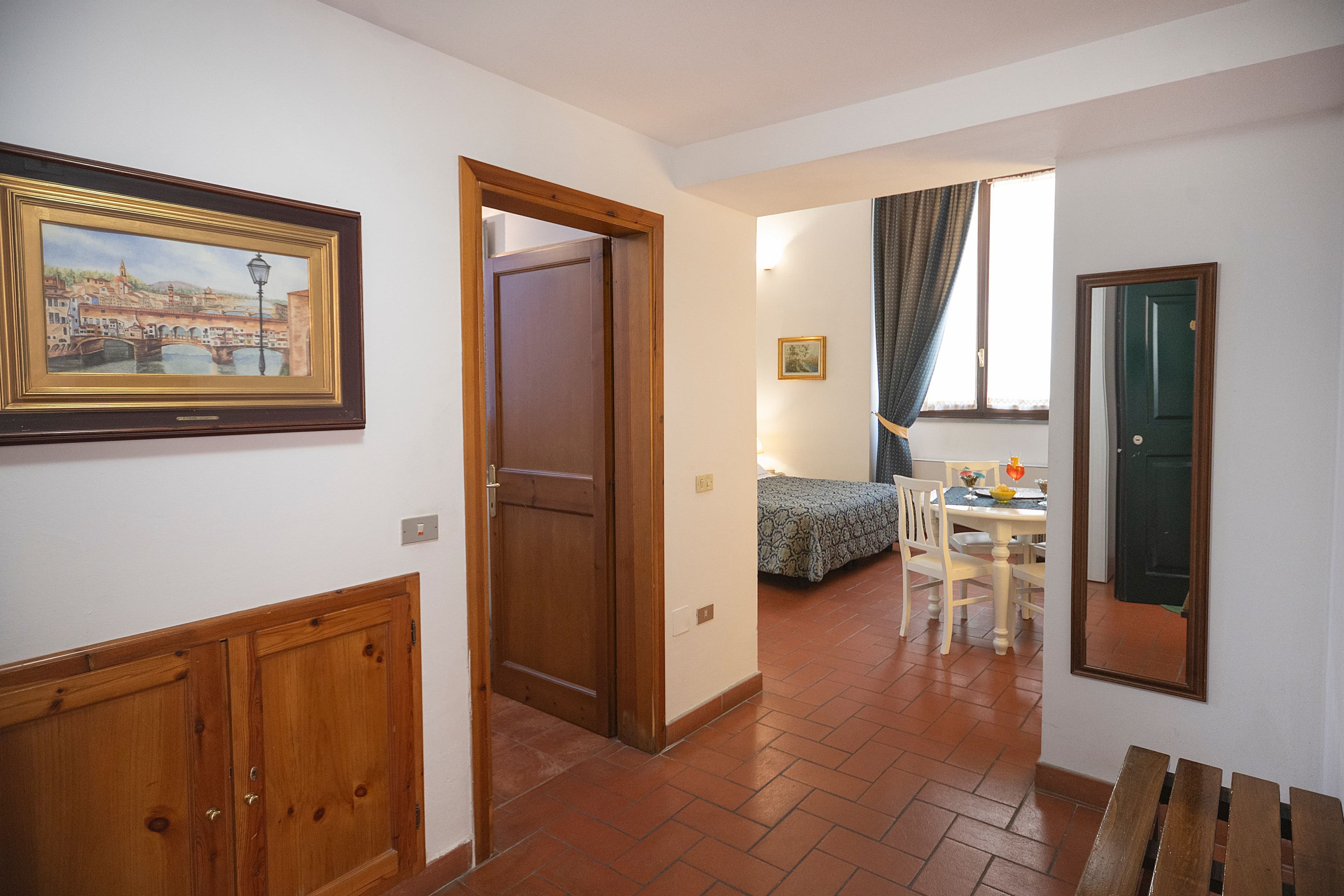 Un pasillo con puerta de madera, un cuadro enmarco, una cama con colcha estampada y una mesa de comedor con sillas.