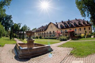 Une cour avec une fontaine, un grand bâtiment doté d’une horloge et un ciel dégagé.