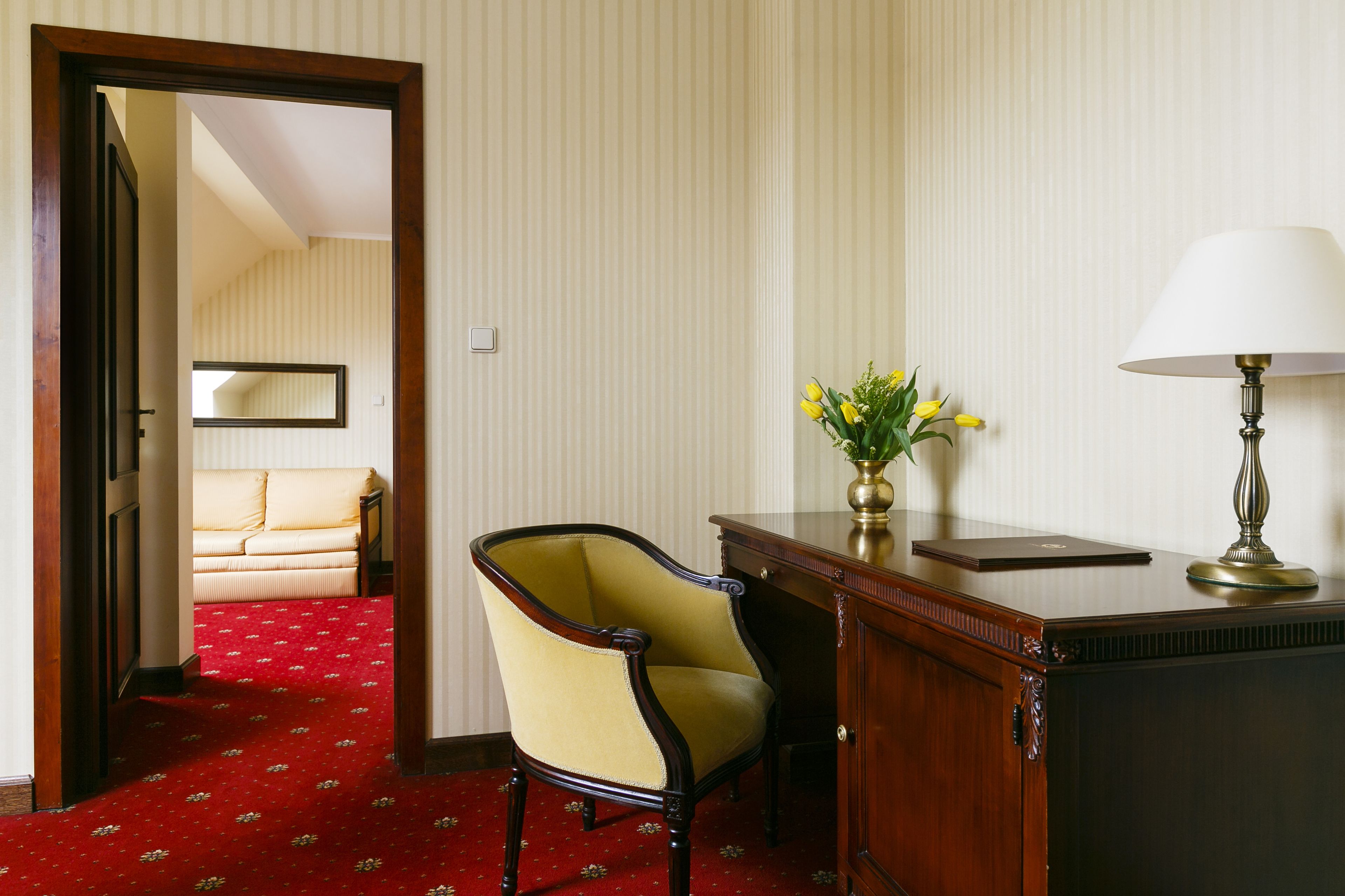 A hotel room with a red carpet, a wooden desk with a lamp, a chair, and a vase with flowers.