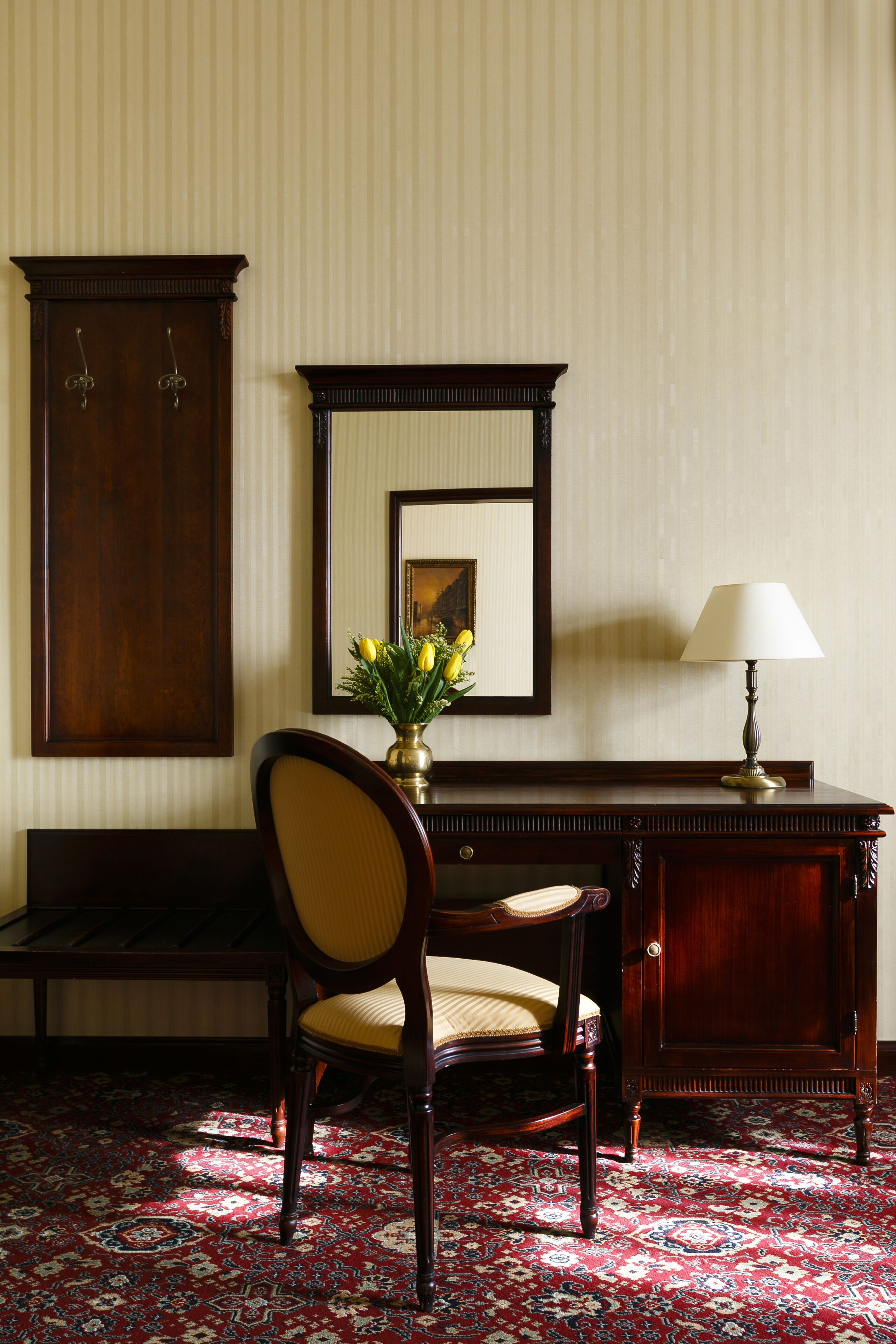 A room with a wooden desk, a chair, a lamp, a mirror, and a vase with flowers.