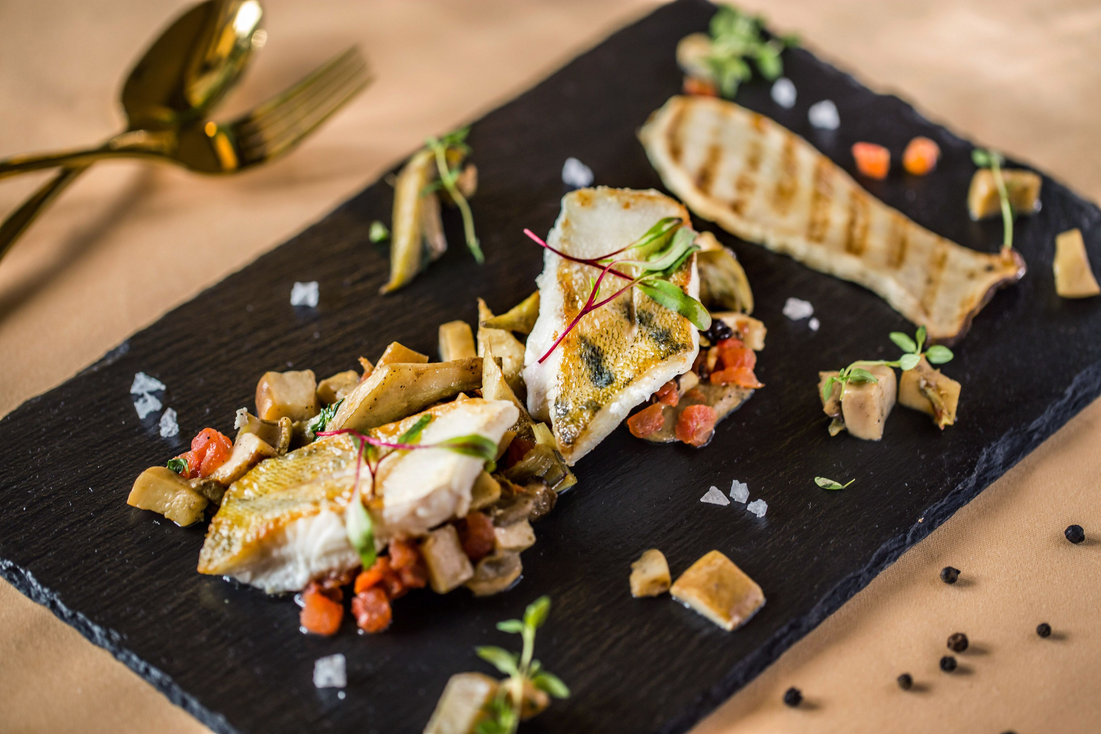 A black slate serving board with a grilled fish fillet, diced potatoes, and a garnish of microgreens.