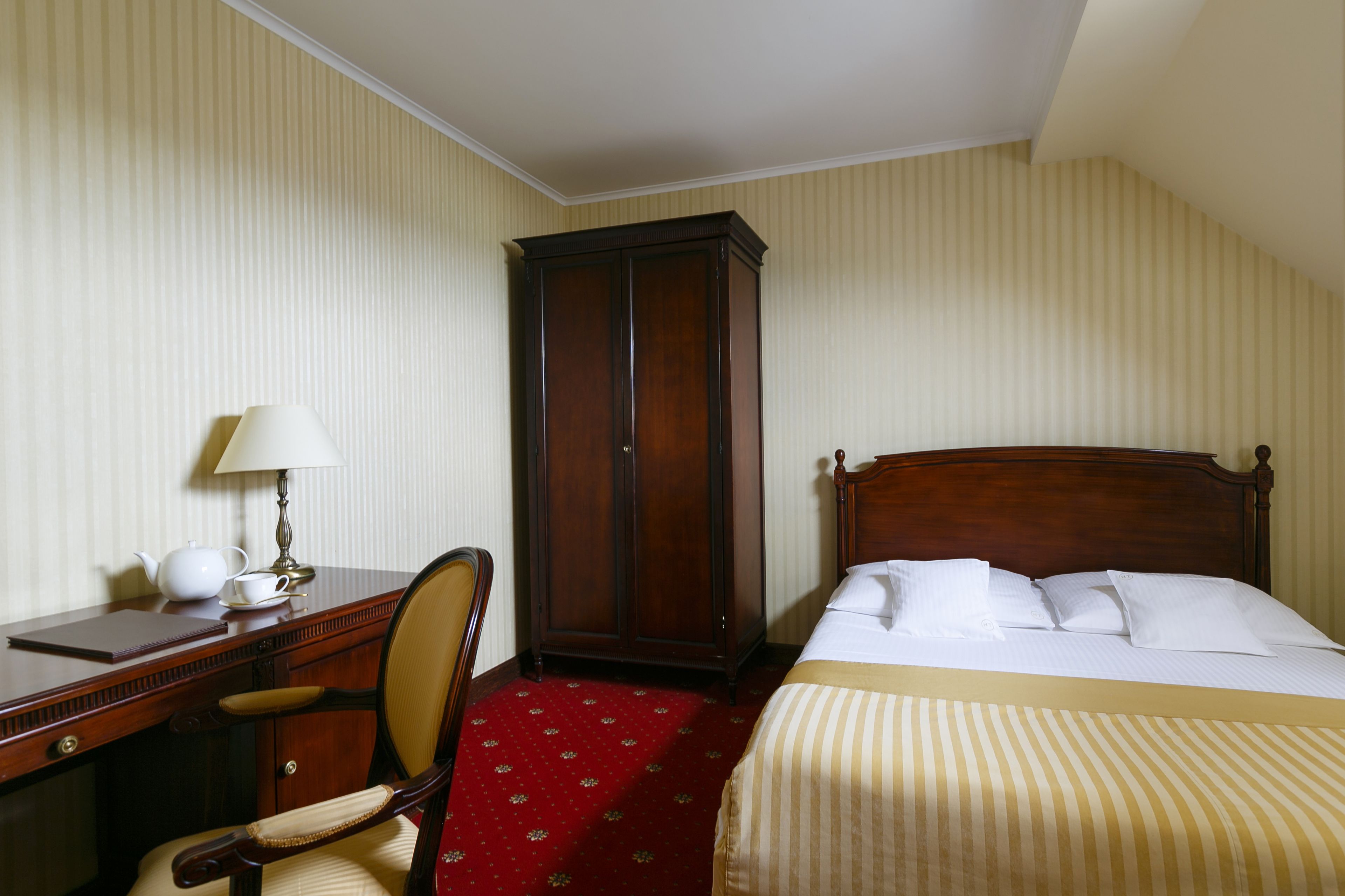 A hotel room with a bed, a desk with a lamp, a chair, and a wardrobe.