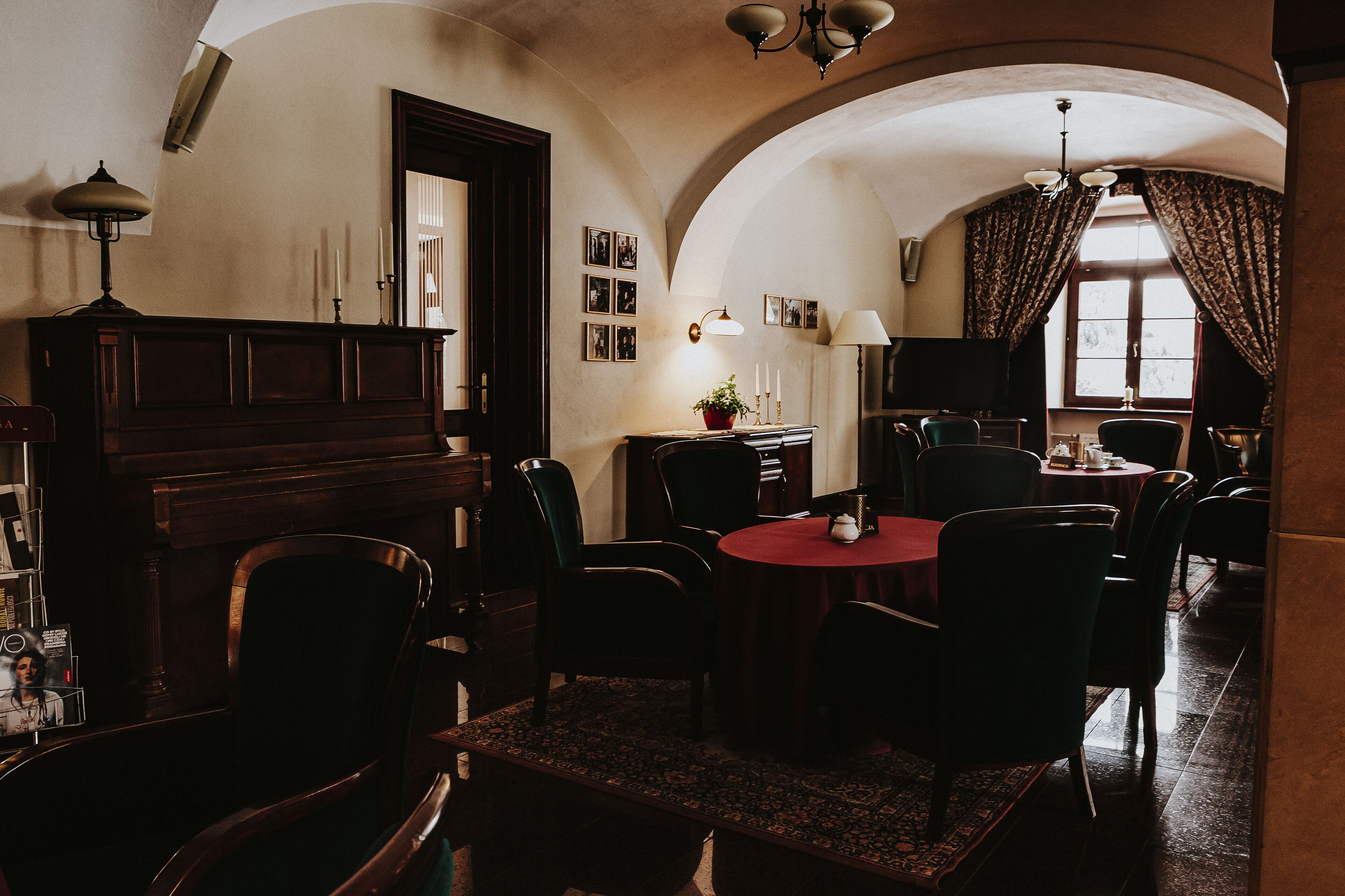 Een eetruimte met een ronde tafel, stoelen en een piano. Er hangen foto's in lijsten aan de muur en een raam met gordijnen.