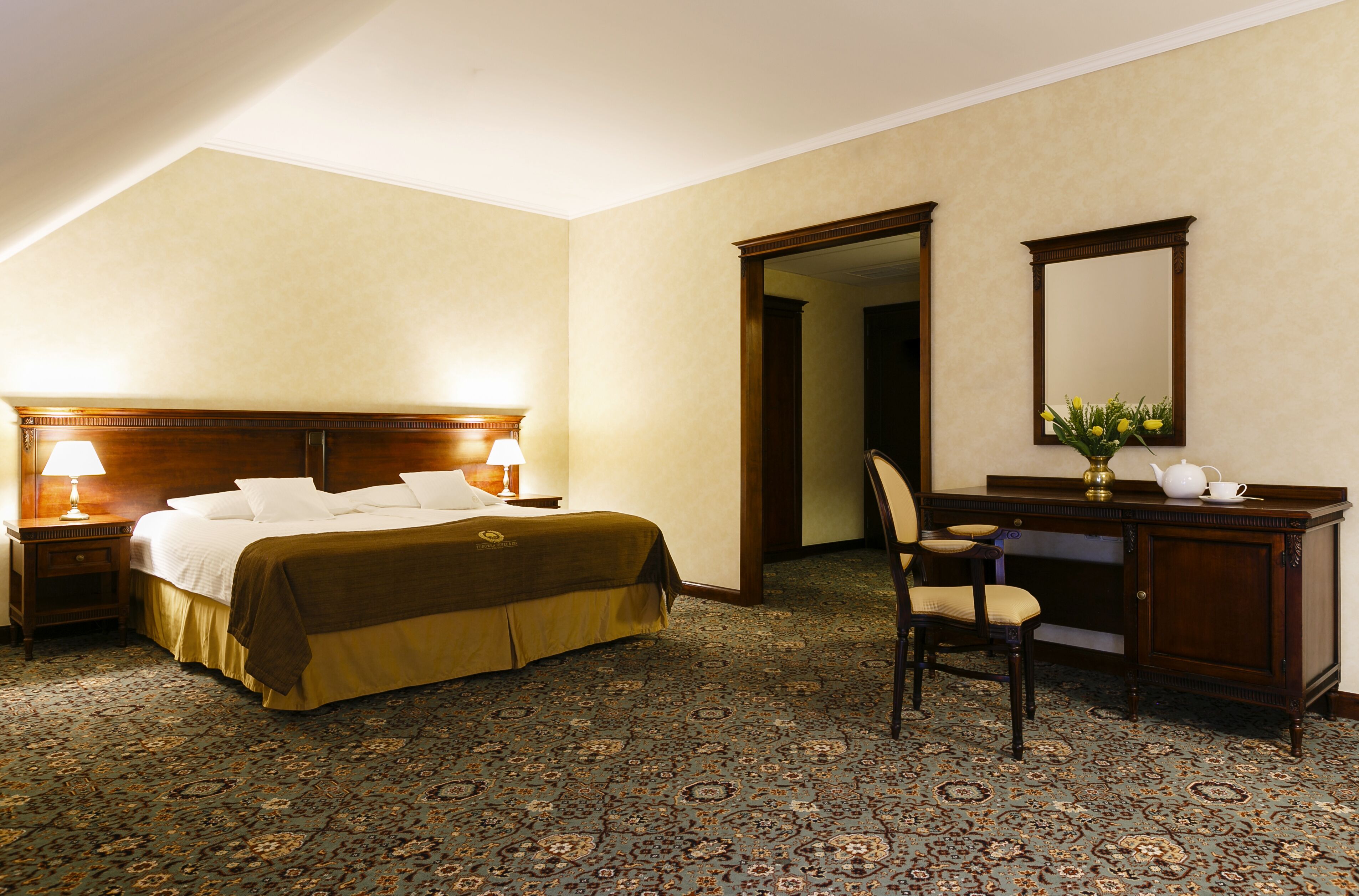 Una habitación de hotel con una cama grande, un escritorio con silla, un espejo y una mesita de noche con lámpara.