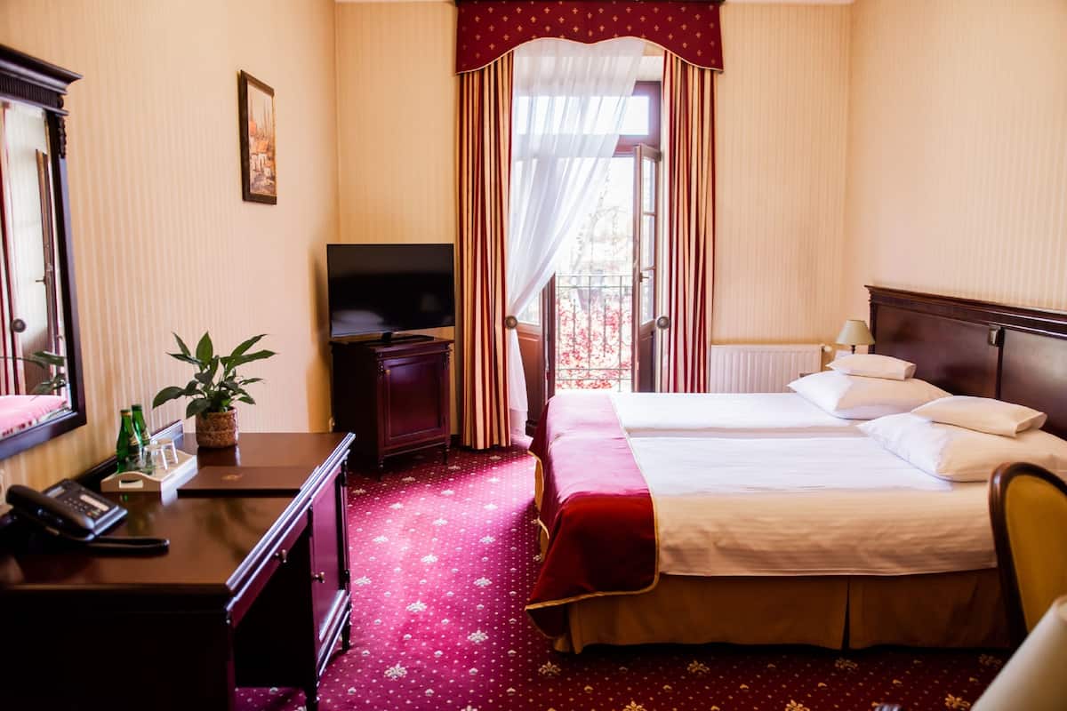 Ein Hotelzimmer mit zwei Betten, einem Fernseher, einem Schreibtisch mit Telefon, einer Pflanze und einem Fenster mit Vorhängen.