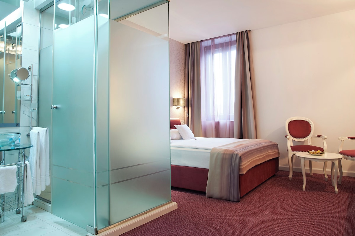 A hotel room with a glass shower enclosure, a bed with a red headboard, a bedside table with a lamp, and a chair with a red cushion.