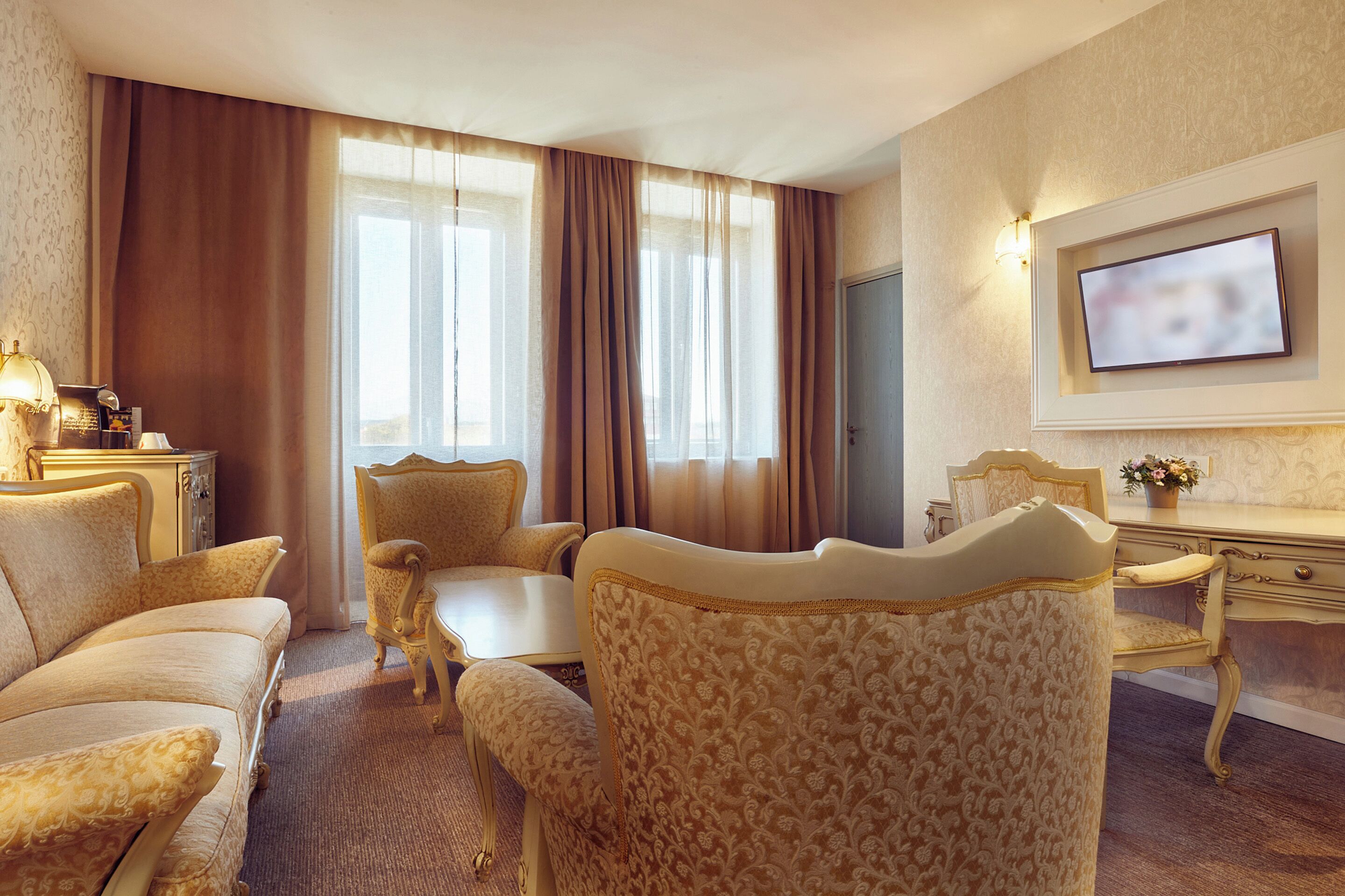 Une chambre d’hôtel avec un canapé, des fauteuils, une petite table, un bureau, une télévision et des rideaux.