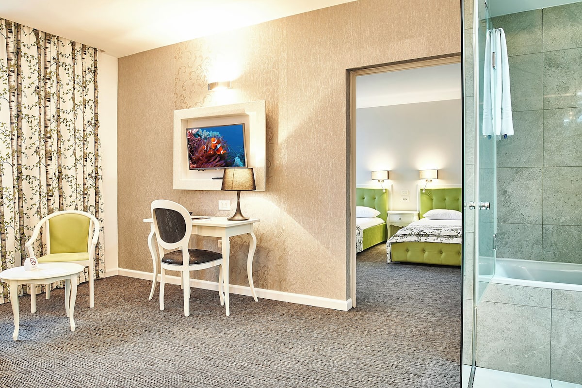 A hotel room with a bed, a desk with a chair, a painting on the wall, and a bathroom with a bathtub.