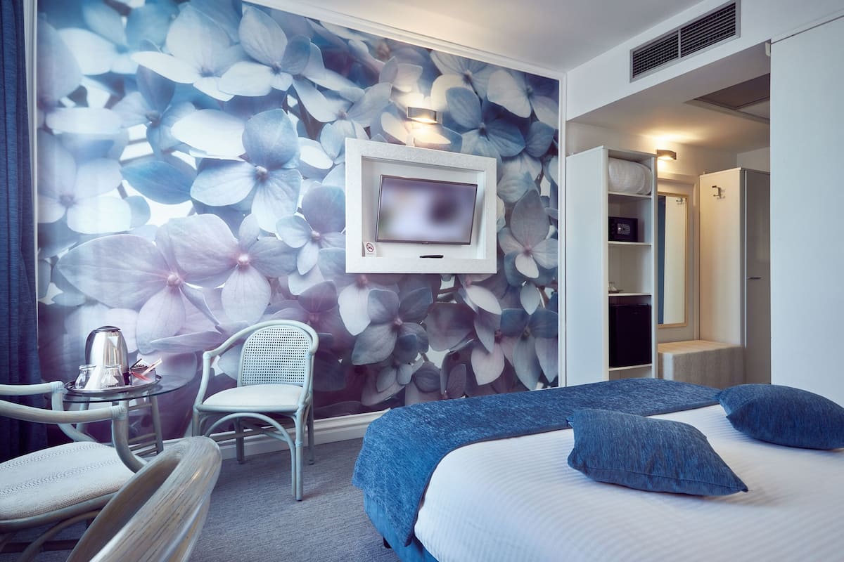 A hotel room with a bed, a TV mounted on the wall, a chair, and a desk with a kettle.