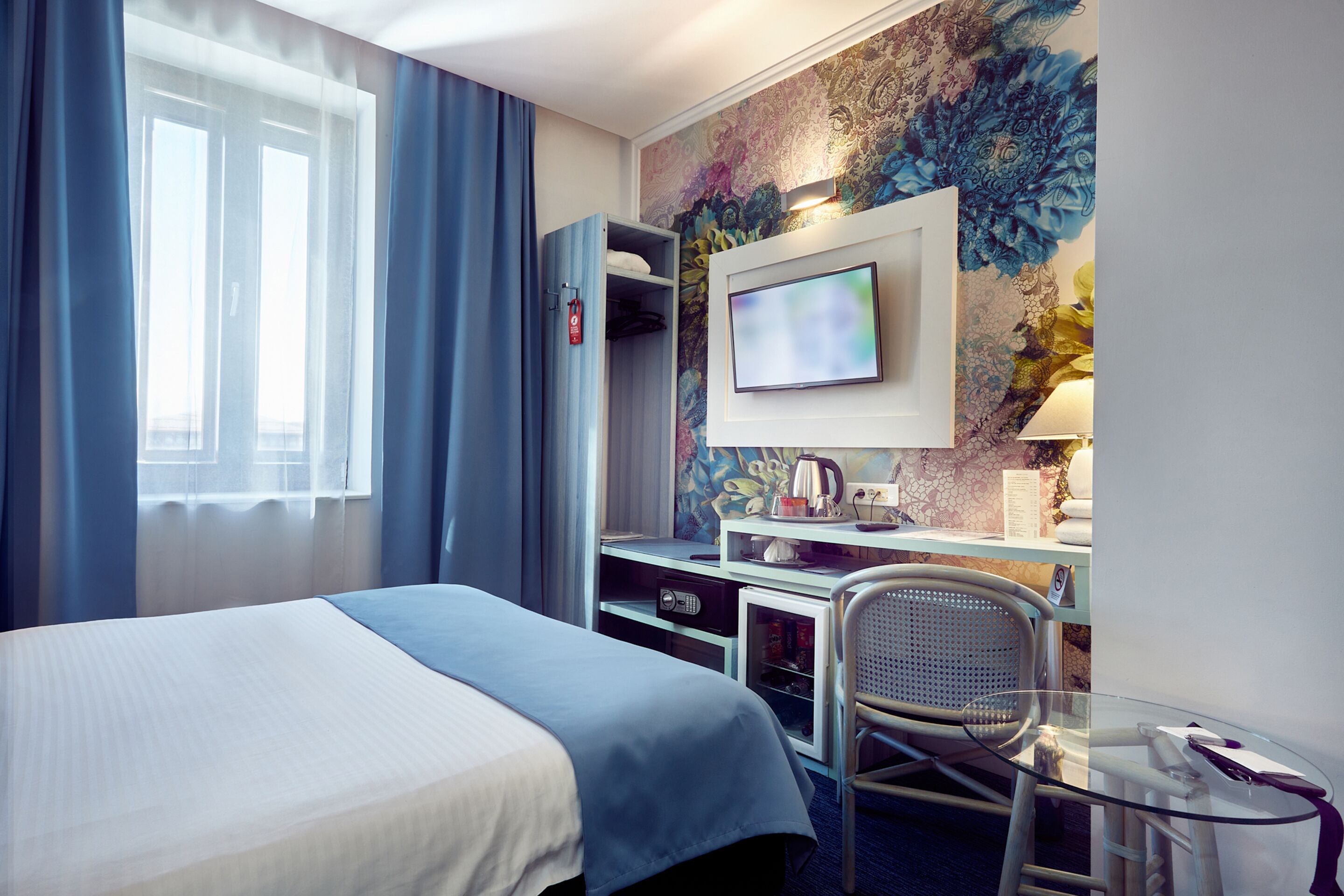 Hotellihuone, jossa on sänky, työpöytä tietokoneella, tuoli, televisio ja ikkuna, jossa on verhot.