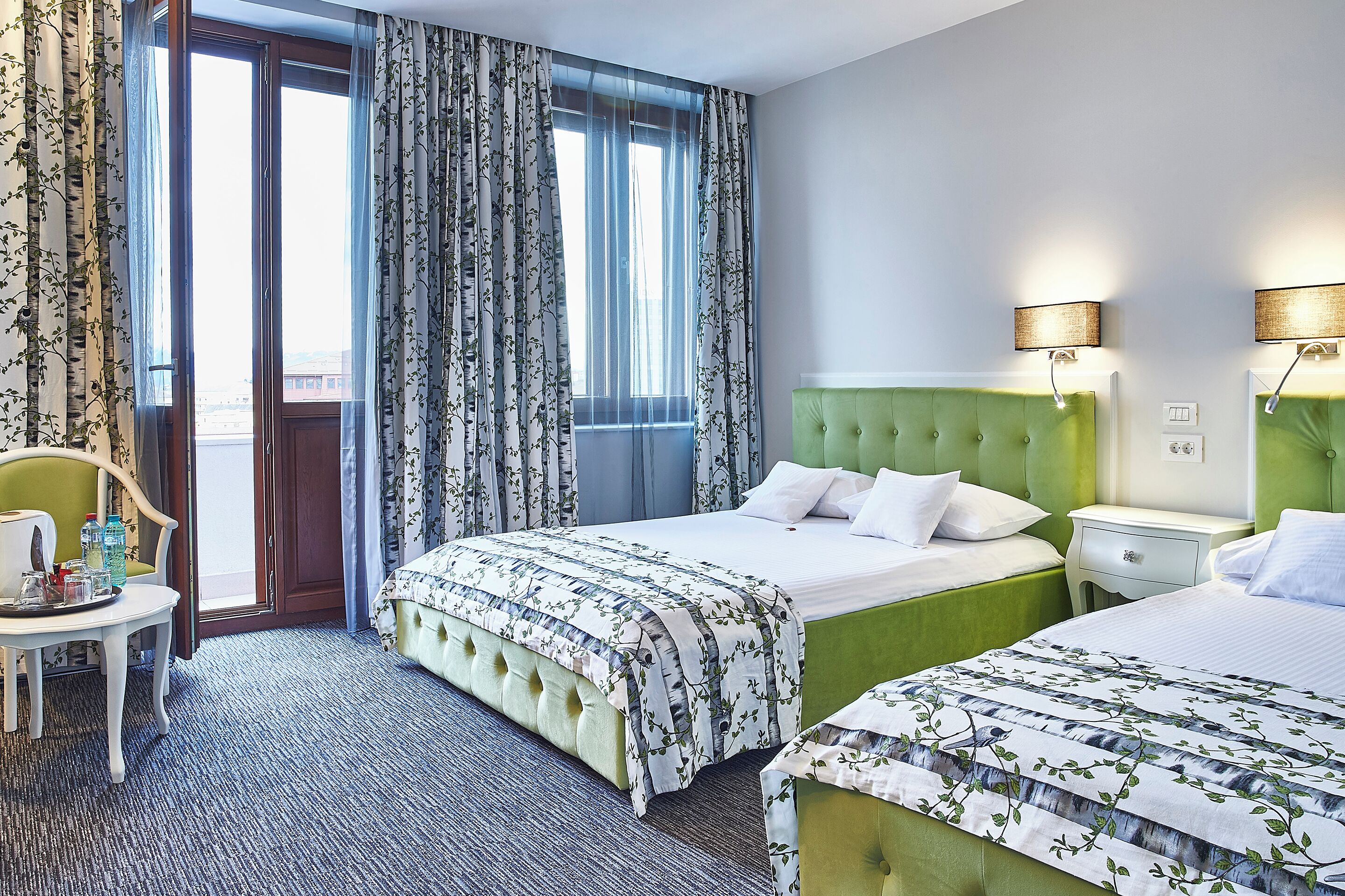 A hotel room with a large bed, a smaller bed, a green upholstered headboard, a bedside table, a chair, and a view through the window.