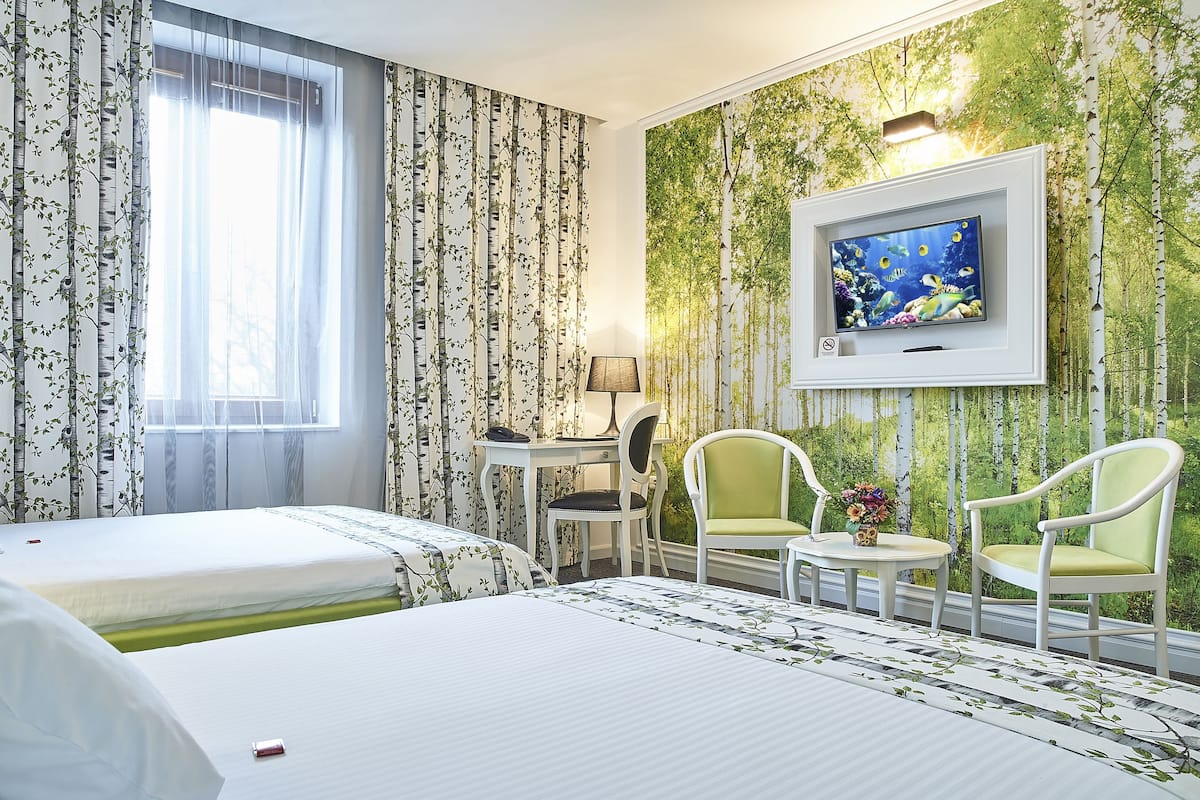 A hotel room with two beds, a desk, two chairs, a TV, and a large window with curtains.