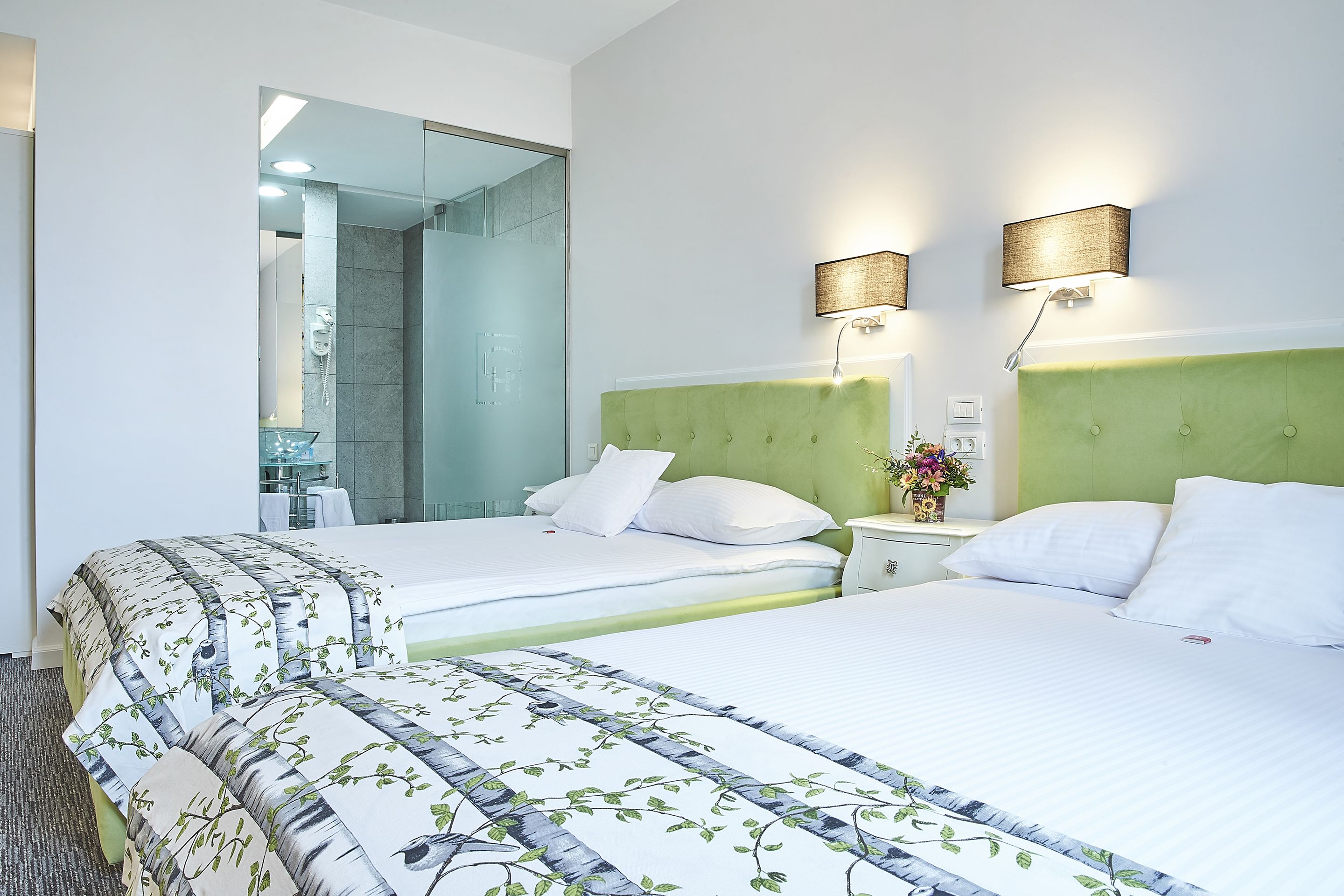 A hotel room with two beds, a green headboard, a nightstand with a vase of flowers, and a bathroom visible through a glass door.
