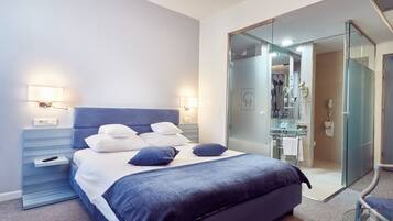 A modern hotel room with a large bed, bedside lamps, a glass-enclosed shower area, and a visible bathroom.