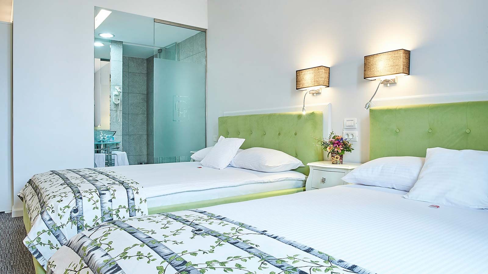 Quarto de hotel com duas camas, cabeceira verde, mesa de cabeceira com um vaso de flores e área de chuveiro com box de vidro.