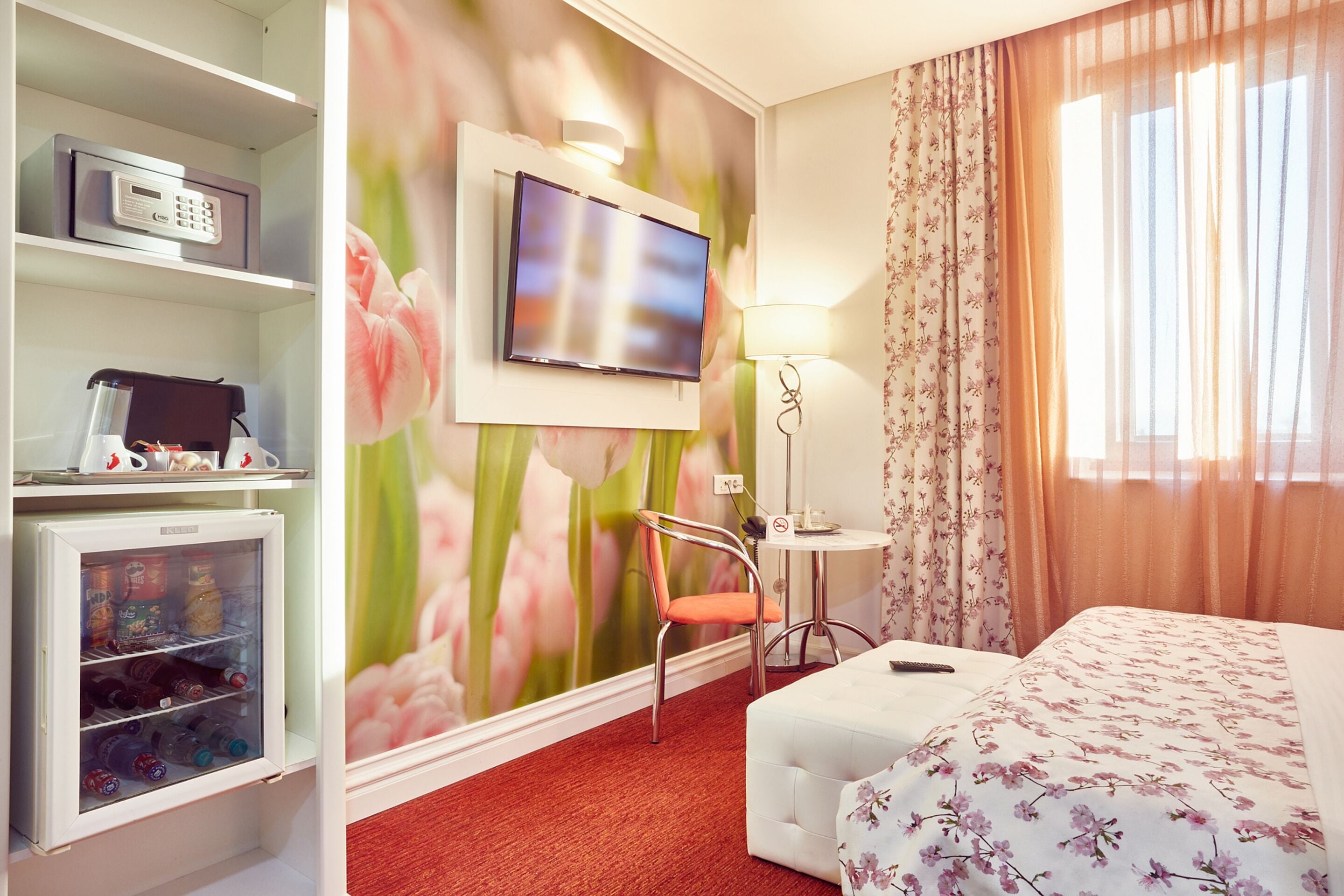 Ein Hotelzimmer mit einem Bett, einem an der Wand befestigten Fernseher, einem kleinen Tisch mit Lampe, einem Kühlschrank und einem Sessel.