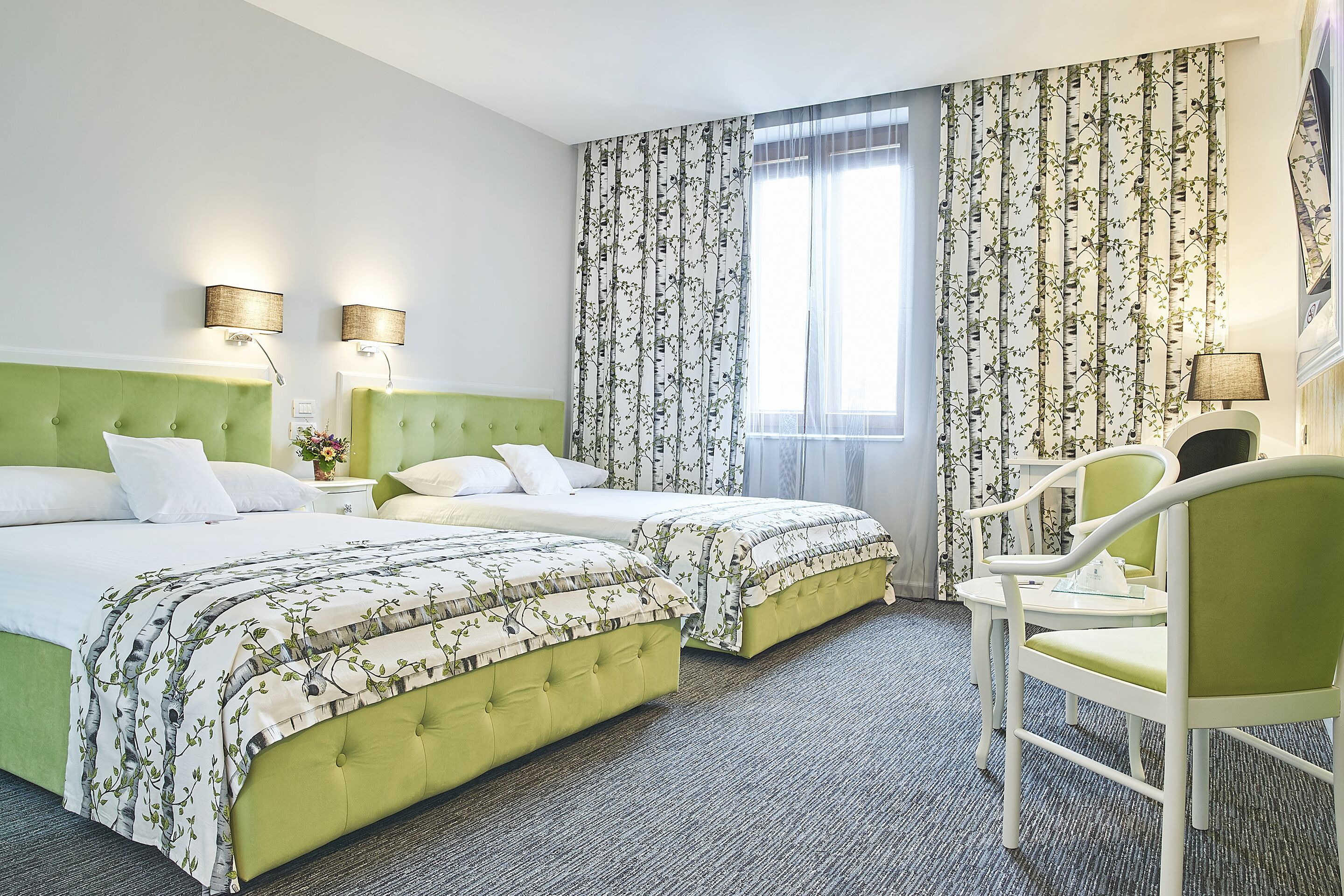 A hotel room with two beds, a green upholstered headboard, a small table with a lamp, and a chair.
