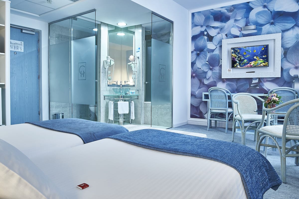 A hotel room with a bed, a bathroom with a glass door, a TV mounted on the wall, and a seating area with chairs.