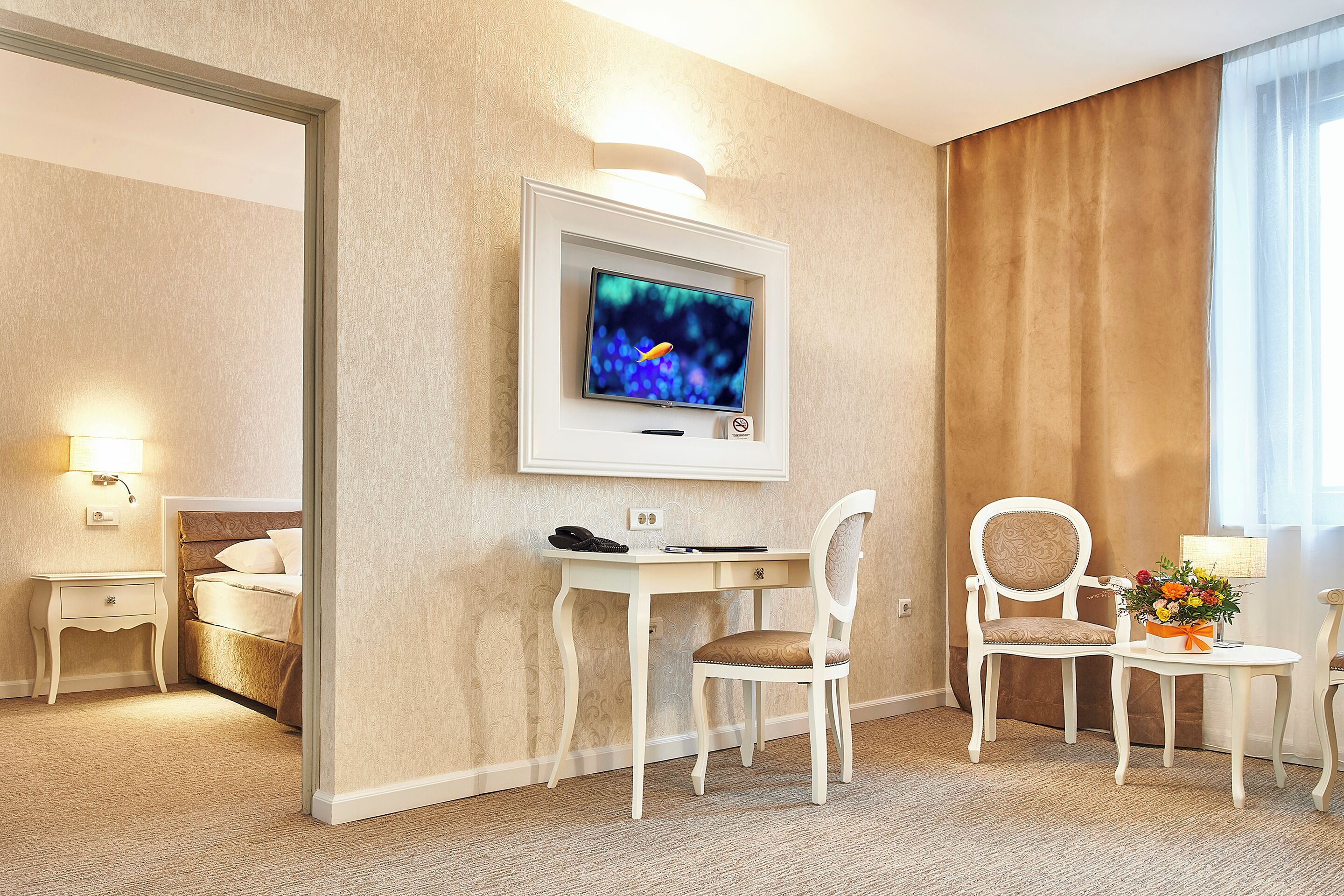 A hotel room with a bed, a desk with a computer, a chair, a small table with a lamp and flowers, and a TV mounted on the wall.