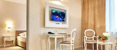 A hotel room with a bed, a desk with a computer, a chair, a small table with a lamp and flowers, and a TV mounted on the wall.