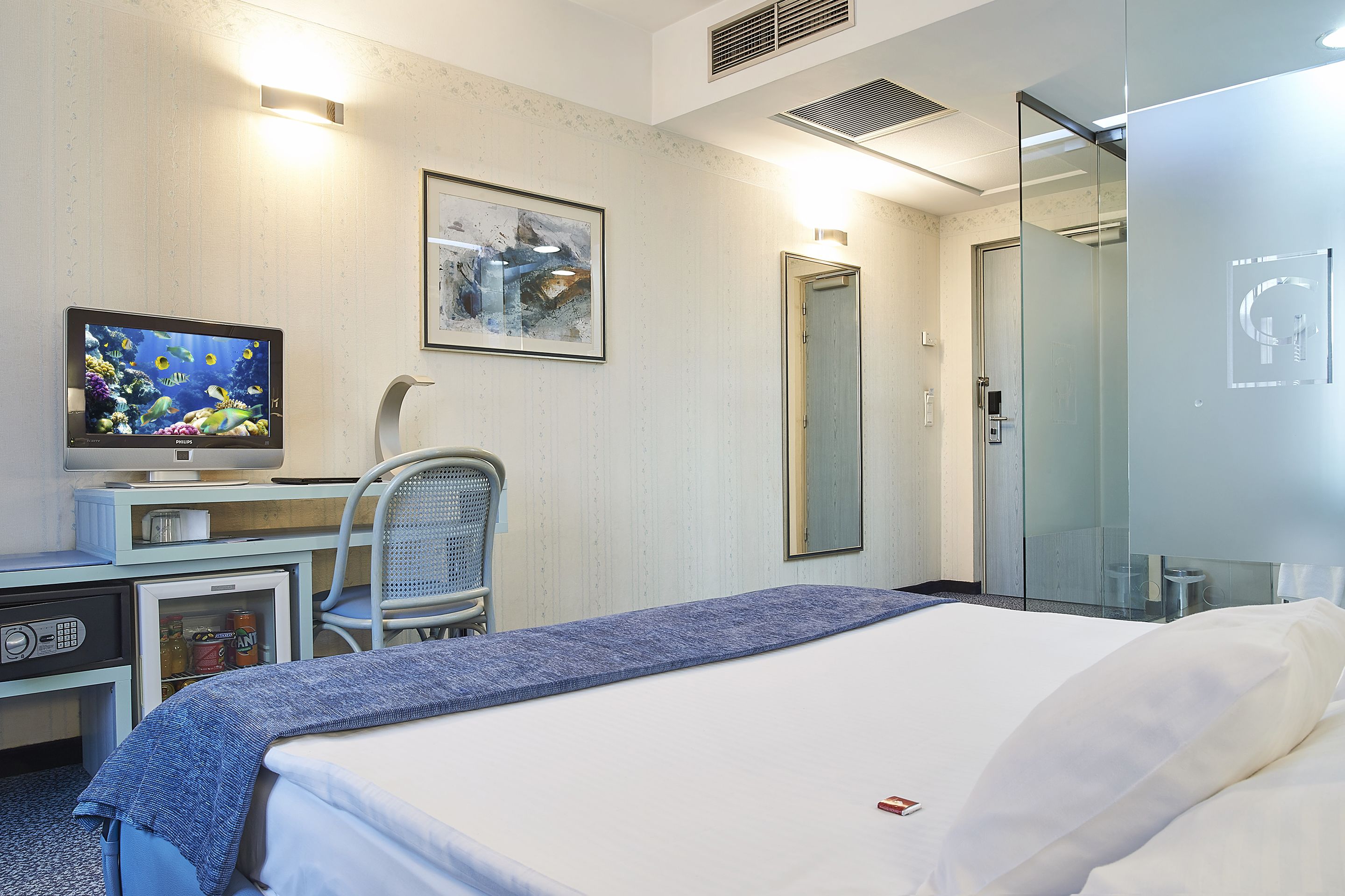 Una habitación de hotel con una cama, un escritorio con computadora, una silla, un televisor y un cuadro en la pared.
