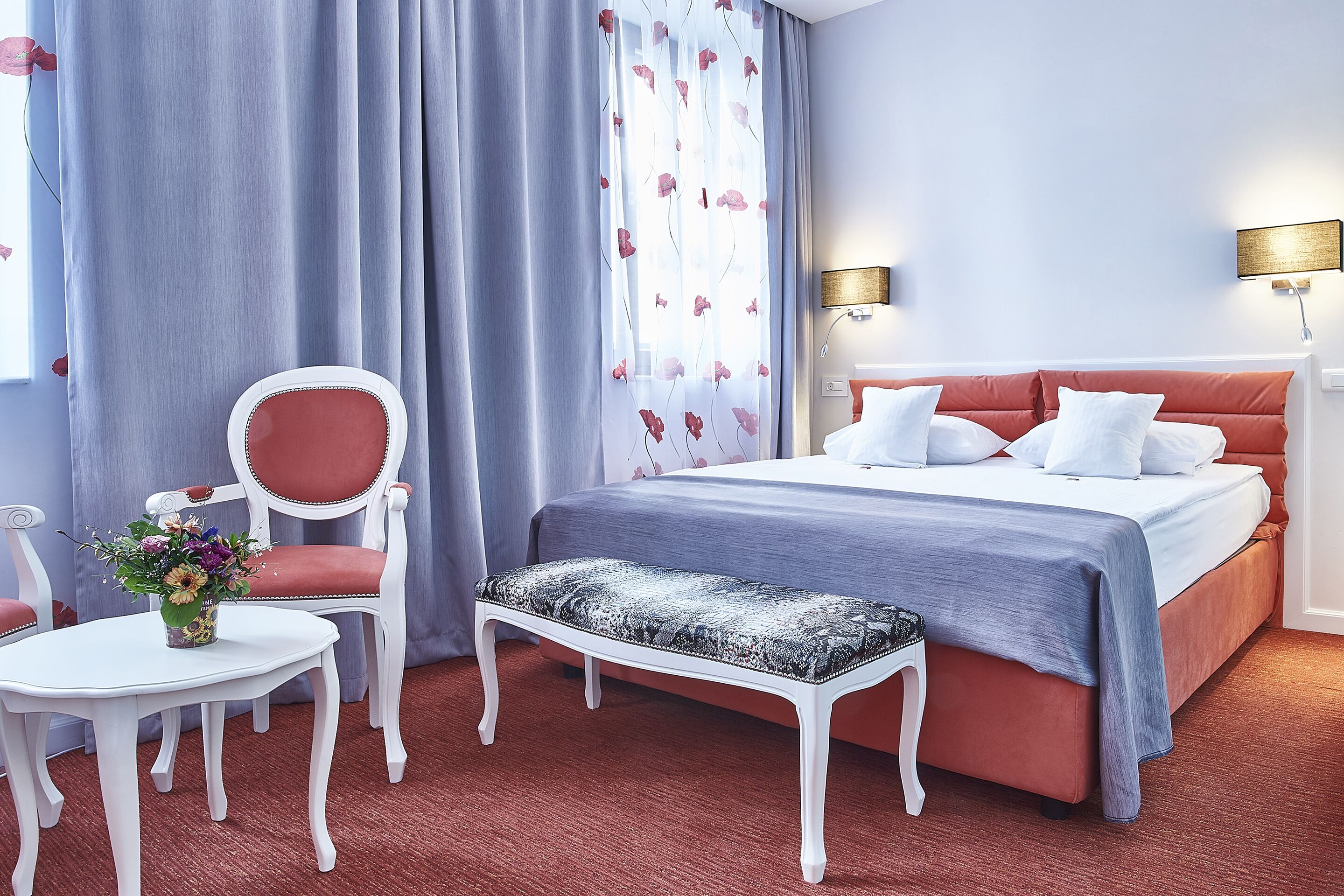 A hotel room with a bed, a chair, a small table, and a bench.