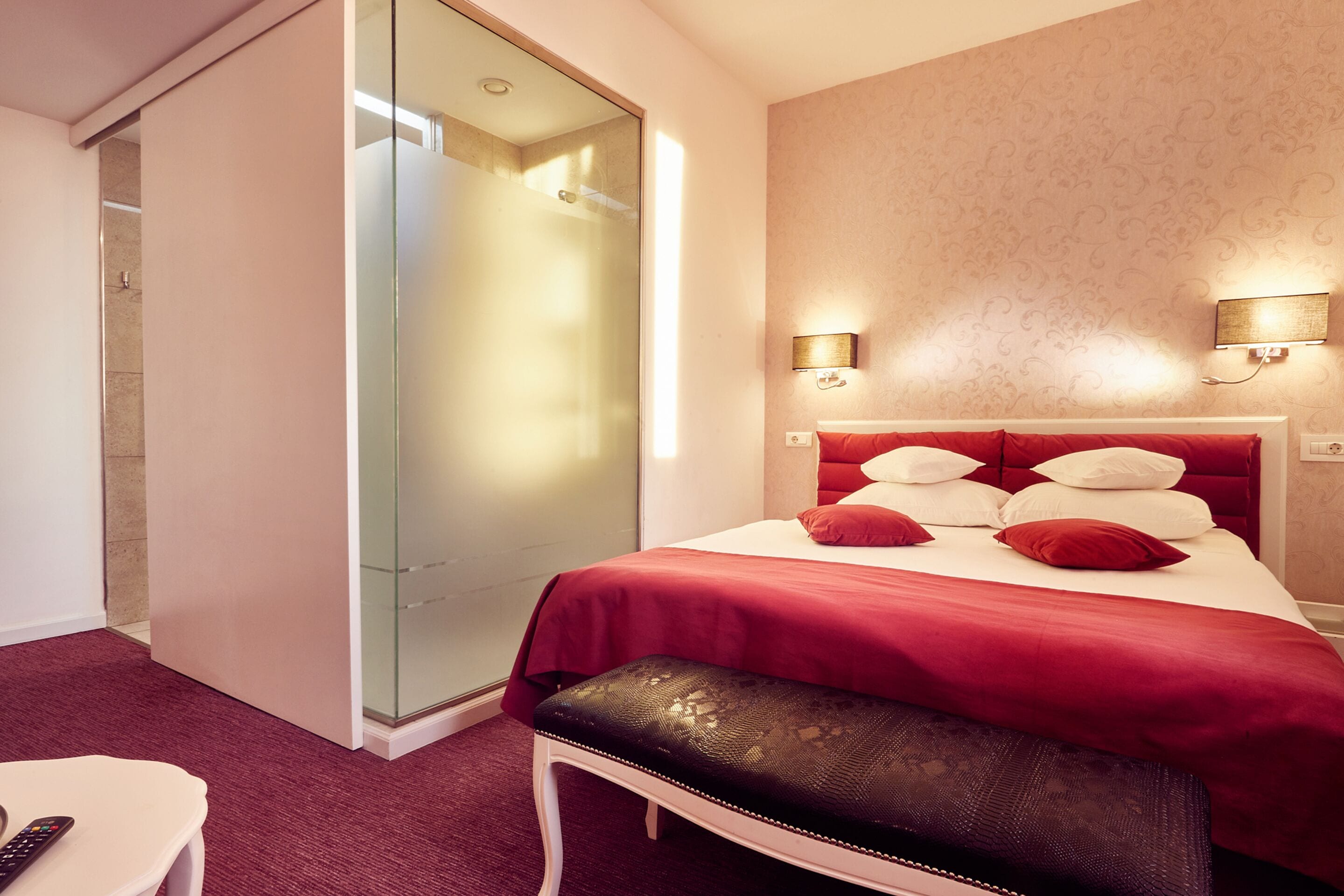 A hotel room with a bed, a bench, a glass shower enclosure, and two wall-mounted lights.