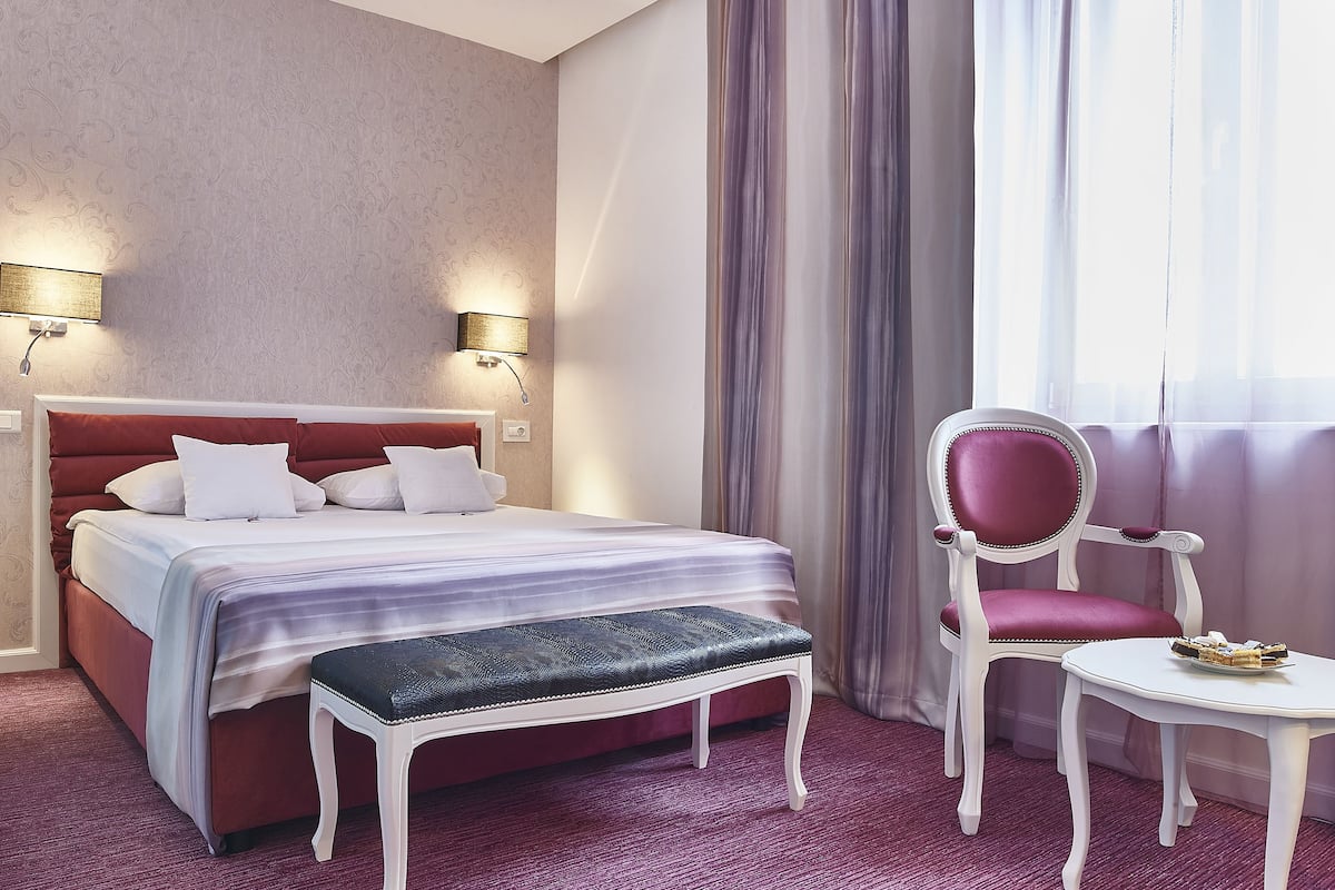 A hotel room with a bed, a bench, a chair, a small table, and two wall-mounted lamps.