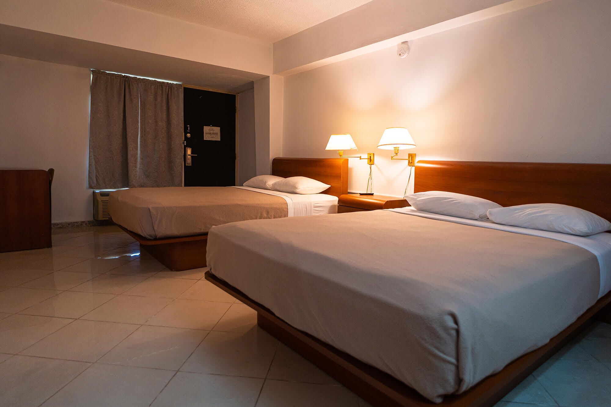 A hotel room with two beds, a wooden headboard, a nightstand with lamps, a door, and a curtain.