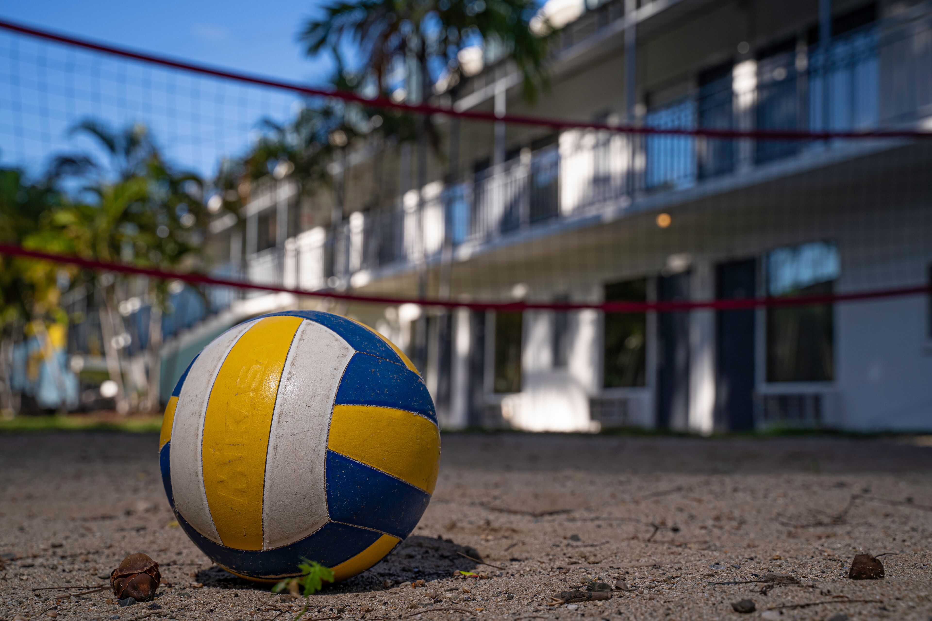 Un voleibol sobre arena, con una red roja al frente y un edificio con balcones en el fondo.