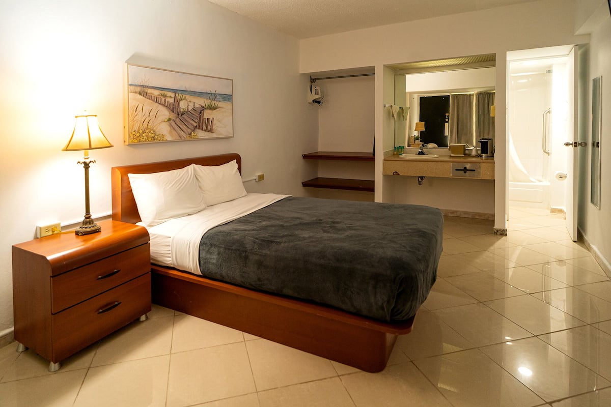 Une chambre à coucher avec un lit, une table de chevet, une lampe et un tableau représentant une scène de plage.
