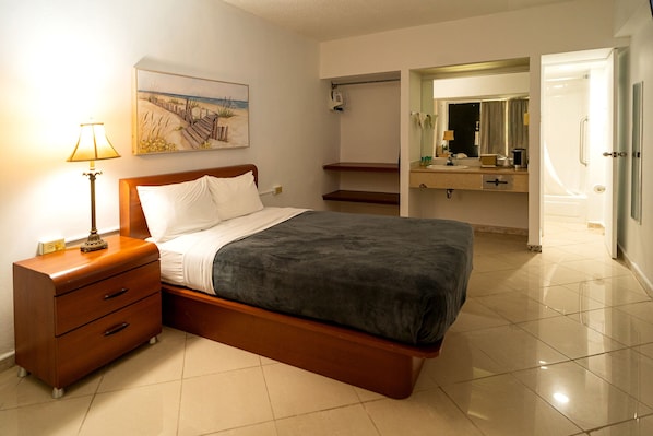 Un dormitorio con cama, mesita de noche, lámpara y un cuadro de una escena de playa.