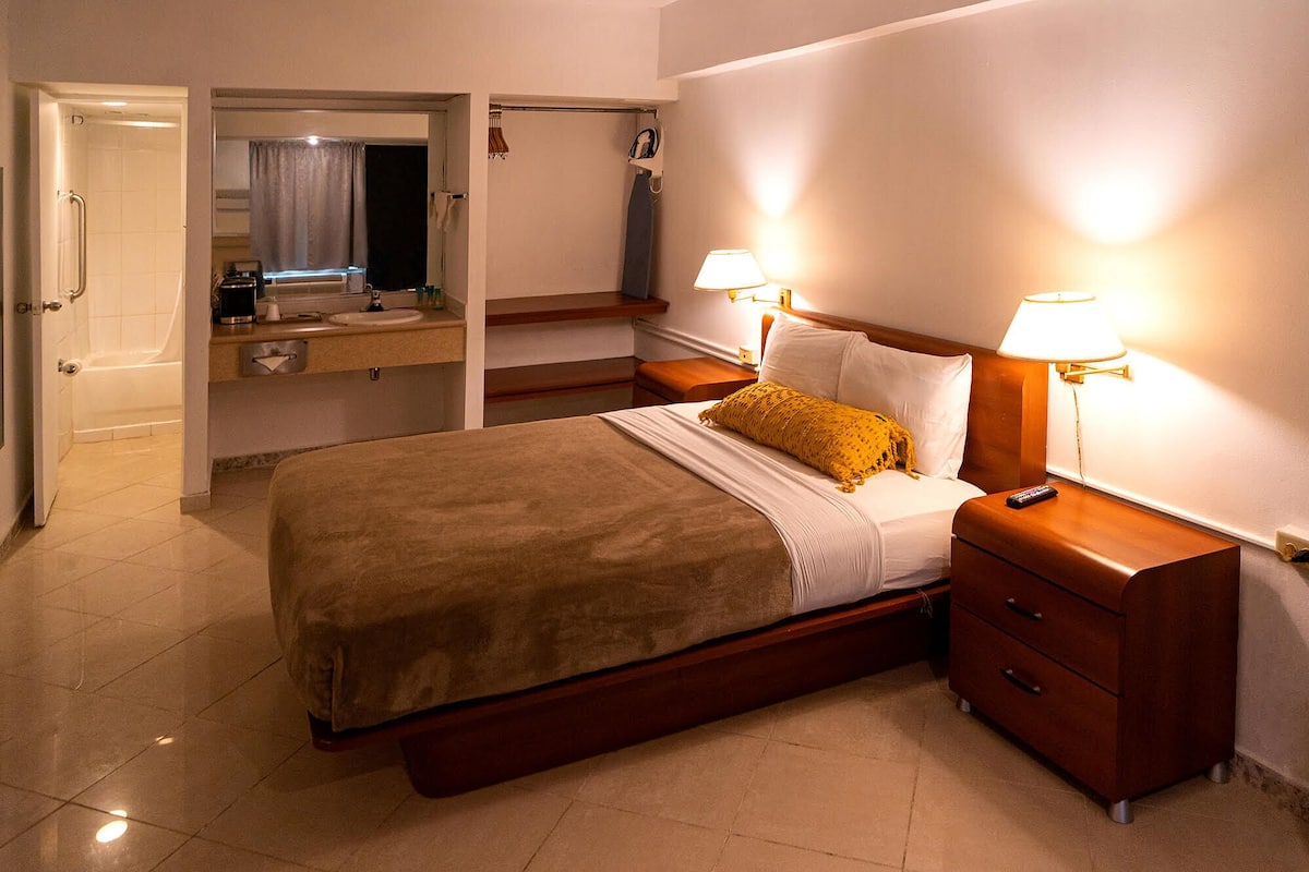 Une chambre d’hôtel comprenant un lit, des tables de chevet, une commode, un miroir, une salle de bain avec douche et un porte-serviettes.