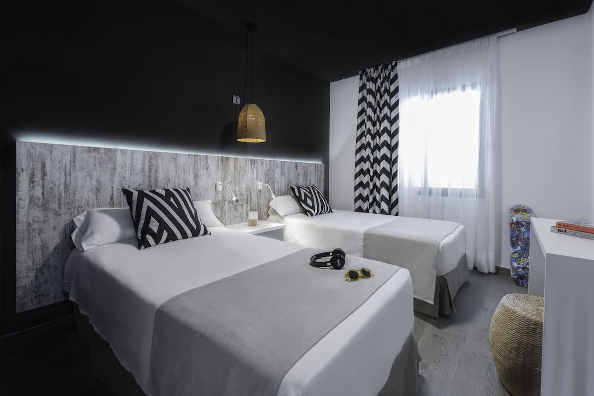 A modern hotel room with two beds, a large window with curtains, a headboard with a light fixture, and a small desk with a chair.