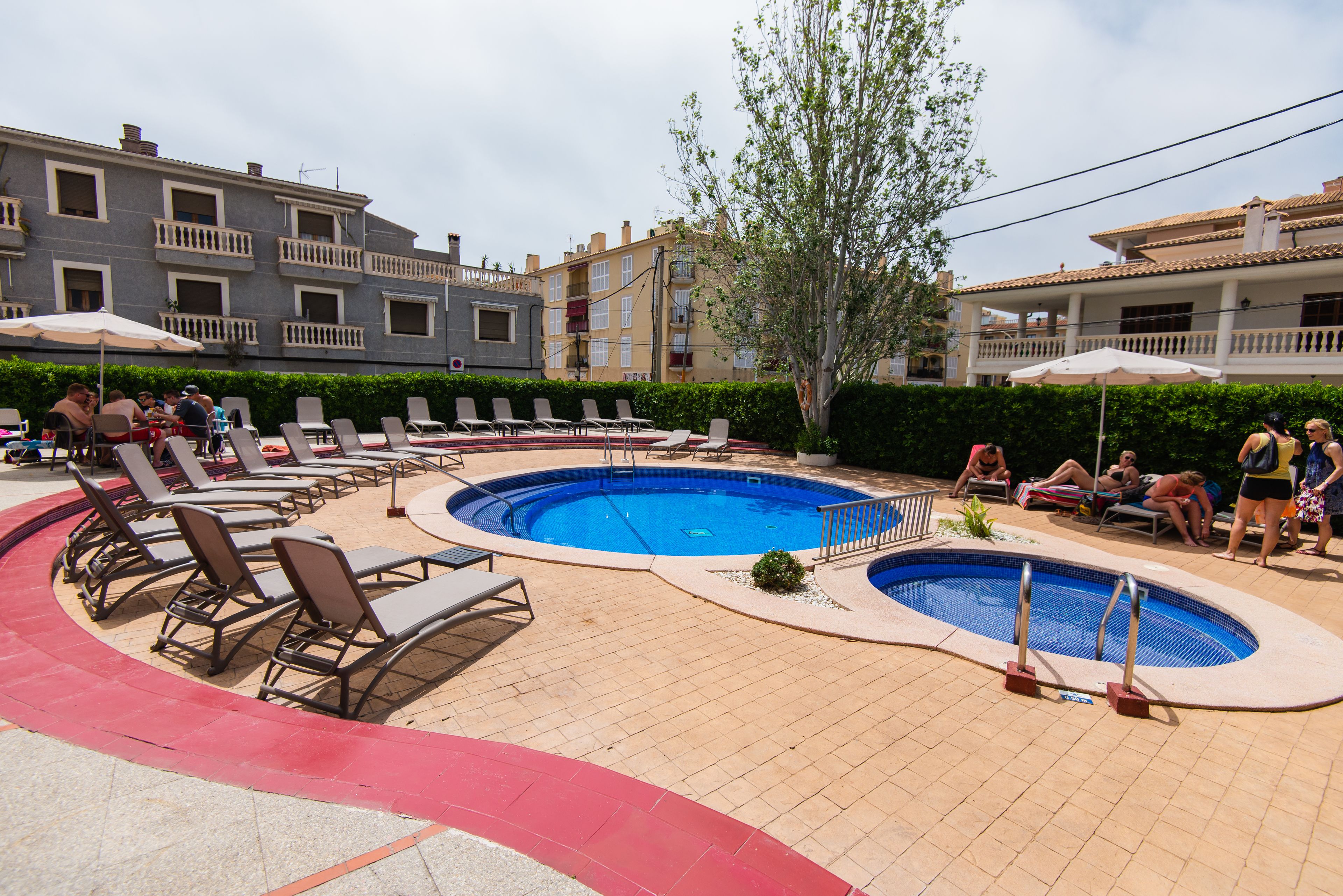Zona de piscina con sillas de descanso y sombrillas, rodeada de edificios.
