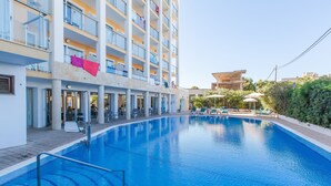 Una piscina al costat d'un hotel de diversos pisos amb balcons, mobiliari exterior i un cel serè.