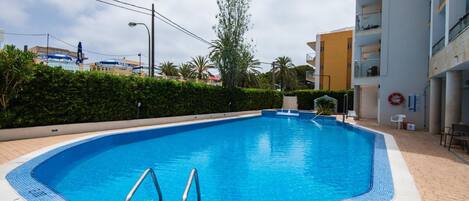 A swimming pool with a tiled edge, surrounded by a hedge and a building with balconies.