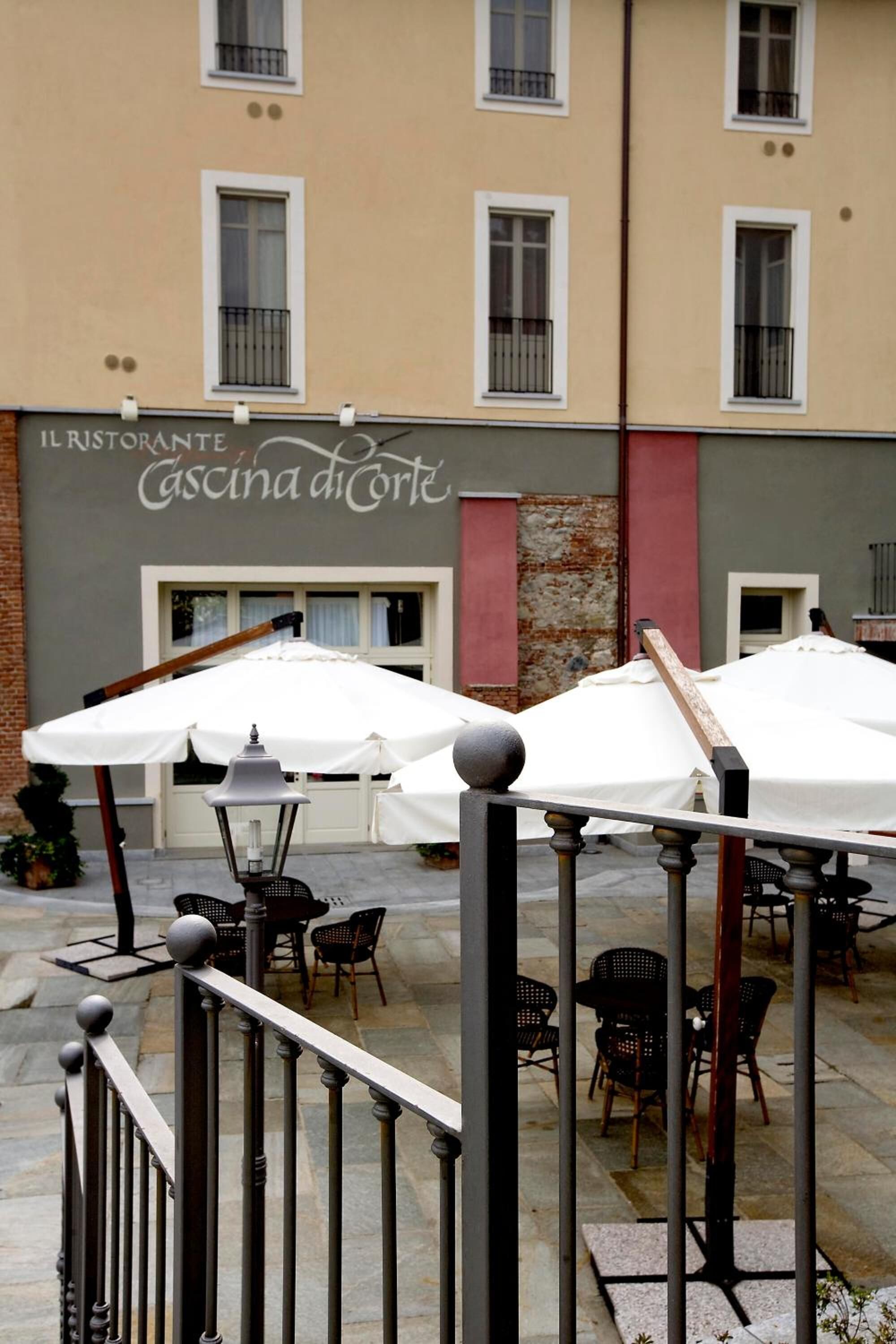 Un espace extérieur aménagé avec des parasols blancs et des chaises noires, situé devant un bâtiment sur lequel on peut lire l’inscription « IL RISTORANTE Cascina di Corte ».
