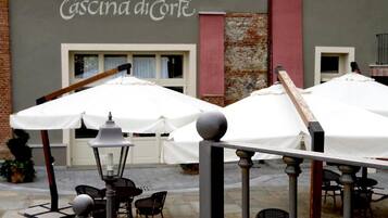 Un espace extérieur aménagé avec des parasols blancs et des chaises noires, situé devant un bâtiment sur lequel on peut lire l’inscription « IL RISTORANTE Cascina di Corte ».