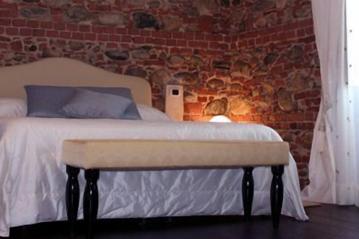 Un dormitorio con pared de piedra, una cama con ropa de cama blanca, un banco y una lámpara fijada a la pared.