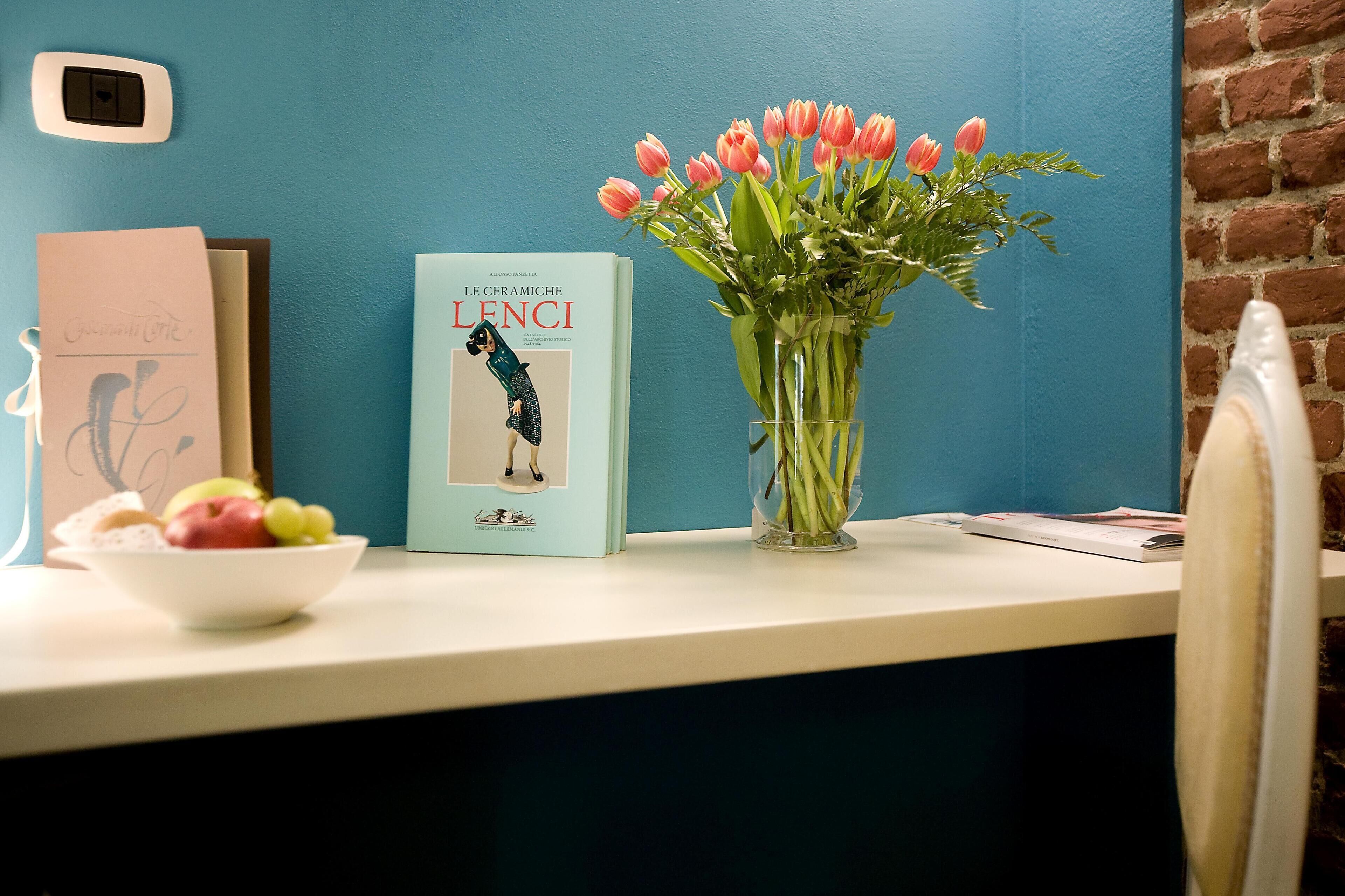 Una mesa con un libro titulado 'LE CERAMICHE LENCI', un jarrón con tulipanes y un frutero con fruta.