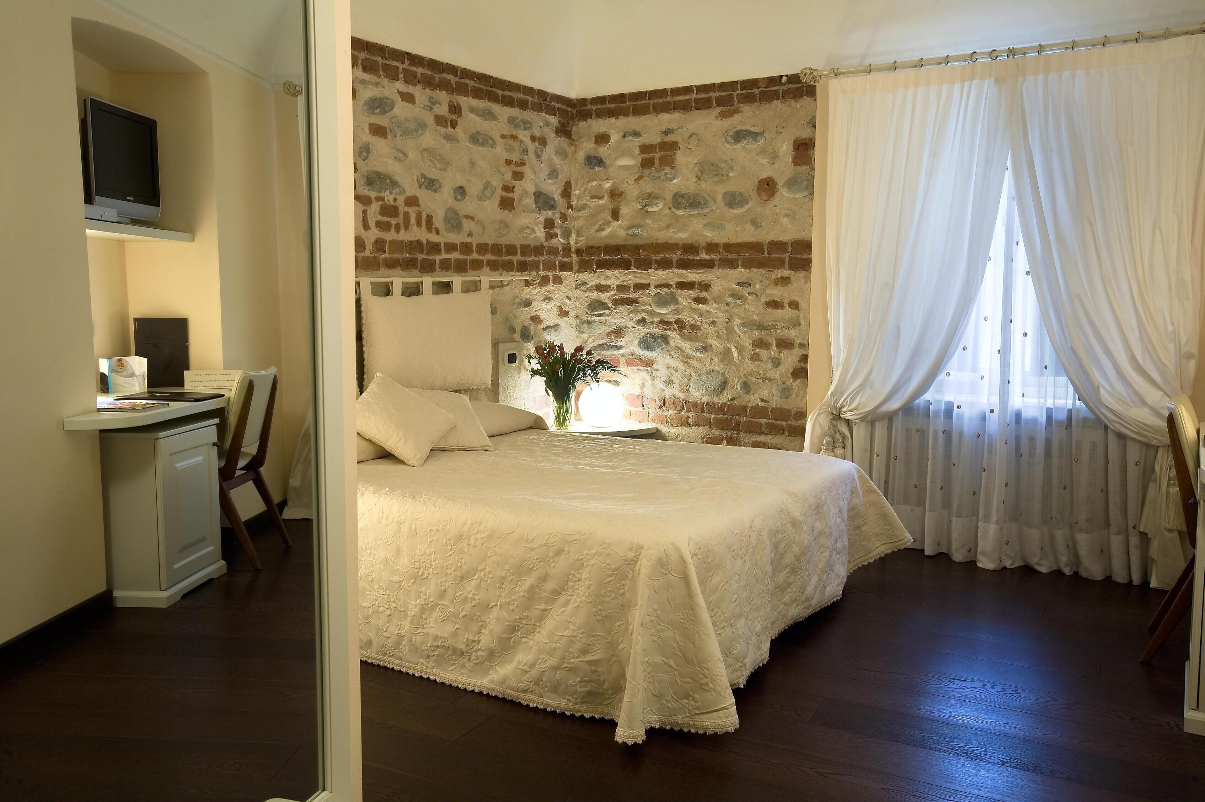 A bedroom with a brick wall, a bed with a white bedspread, a desk with a computer, and a window with curtains.