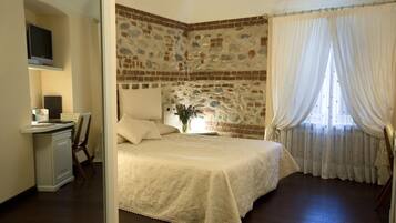 A bedroom with a brick wall, a bed with a white bedspread, a desk with a computer, and a window with curtains.