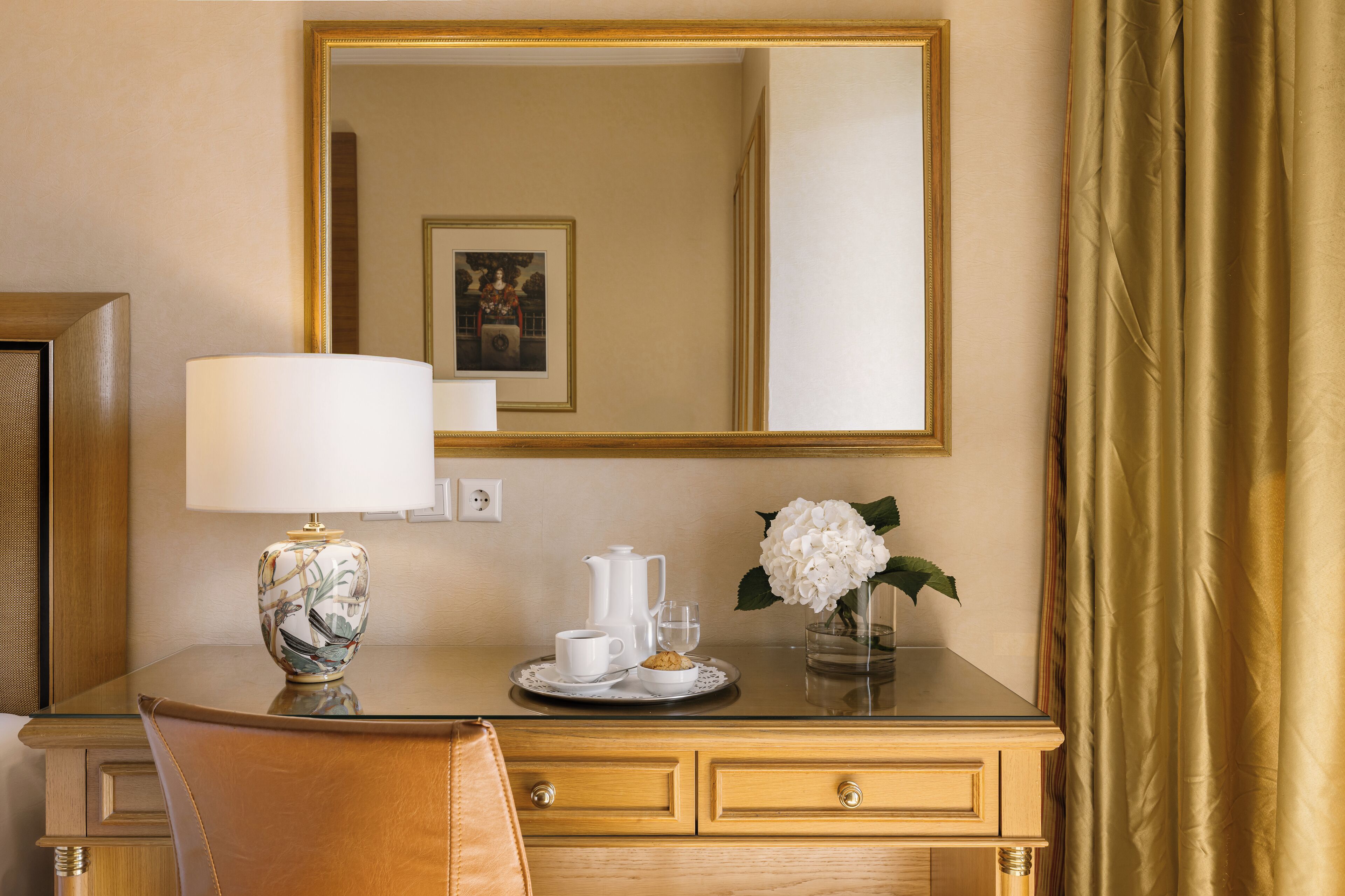 Ein Hotelzimmer mit einem hölzernen Schreibtisch, einer Lampe, einem Tablett mit Teekanne, Tassen und einem Keks. Über dem Schreibtisch befindet sich ein Spiegel und auf der rechten Seite ein goldener Vorhang.