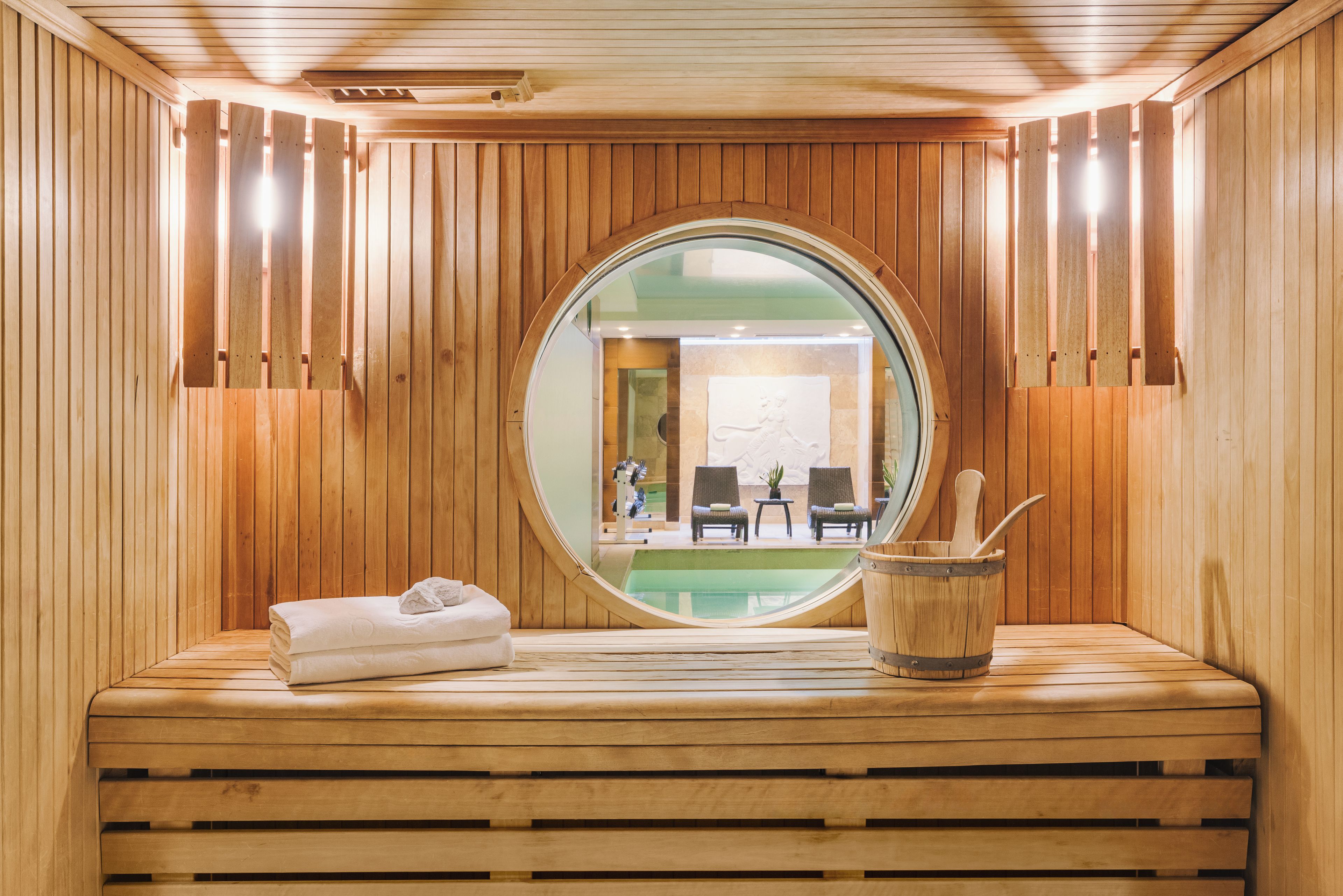 Una sauna con paredes de madera, un espejo redondo, un banco y un cubo con una cuchara.