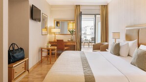 A hotel room with a large bed, a desk with a chair, a small table, a TV, and a view of the outside. - Divani Palace Larissa (Larissa)