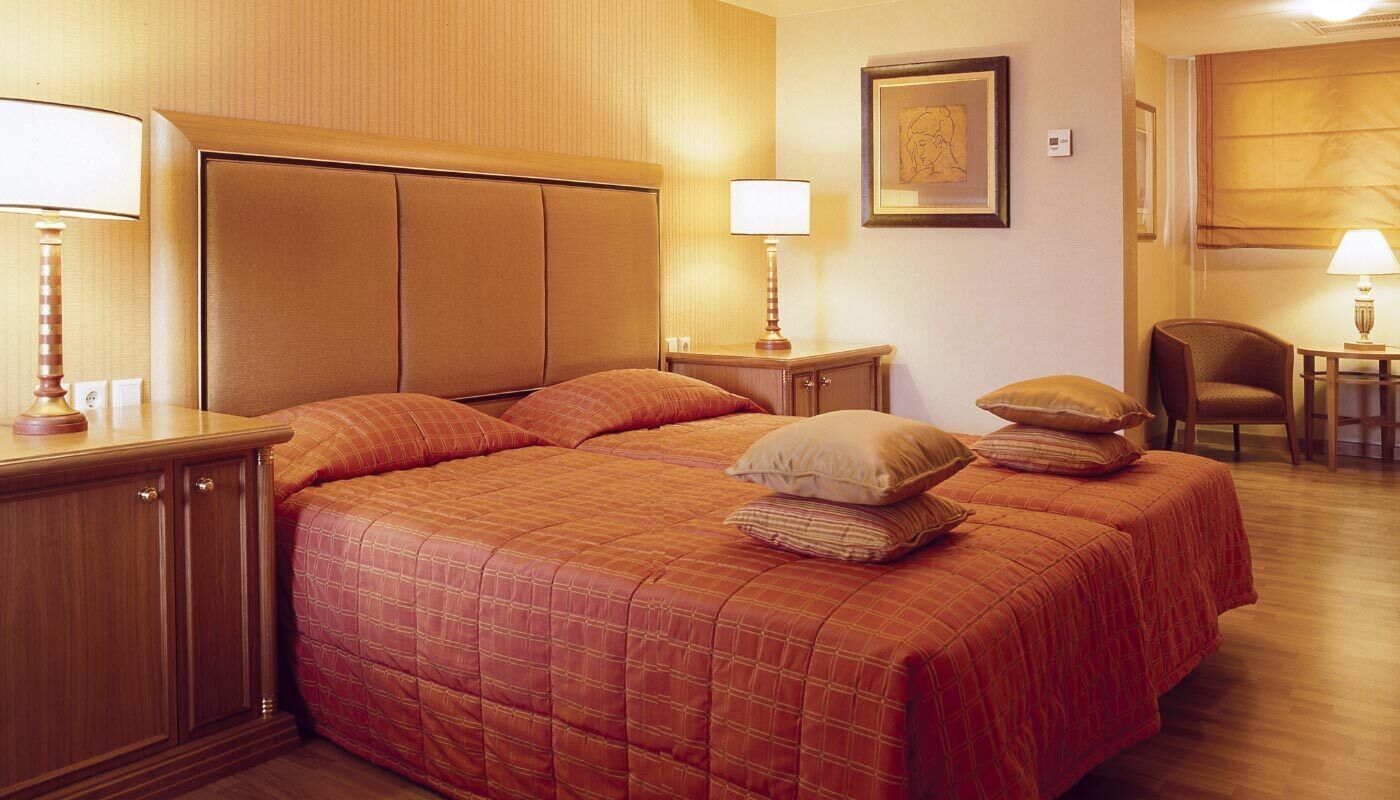A hotel room with a large bed, two bedside tables with lamps, a wooden headboard, a framed artwork on the wall, and a chair in the corner.