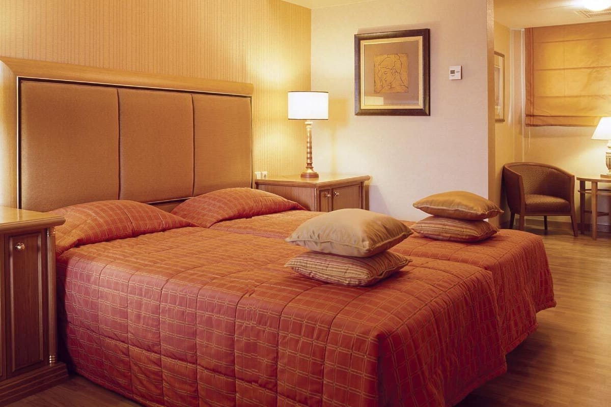 A hotel room with a large bed, two bedside tables with lamps, a wooden headboard, a framed artwork on the wall, and a chair in the corner.